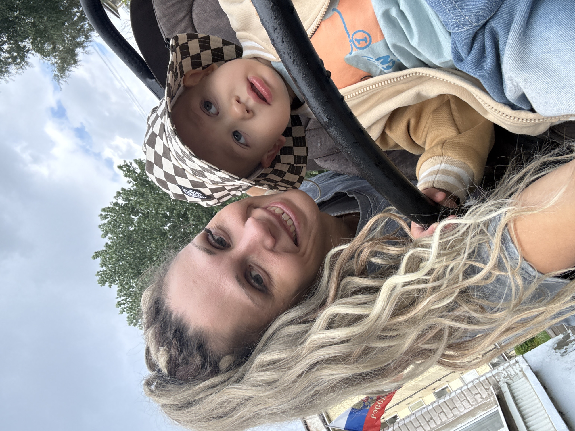A woman with blonde, wavy hair pulled back in a ponytail, wearing a gray shirt, is taking a selfie with a baby. The baby is wearing a checkered bucket hat, a light brown jacket with striped cuffs, and a light blue t-shirt. The baby is in a stroller, and the woman is leaning in from the side to be in the frame. In the background, a Russian flag is visible next to a building with light-colored brickwork. Trees are also visible in the background under a cloudy sky. The location is identified as Rusanovo, Russia.