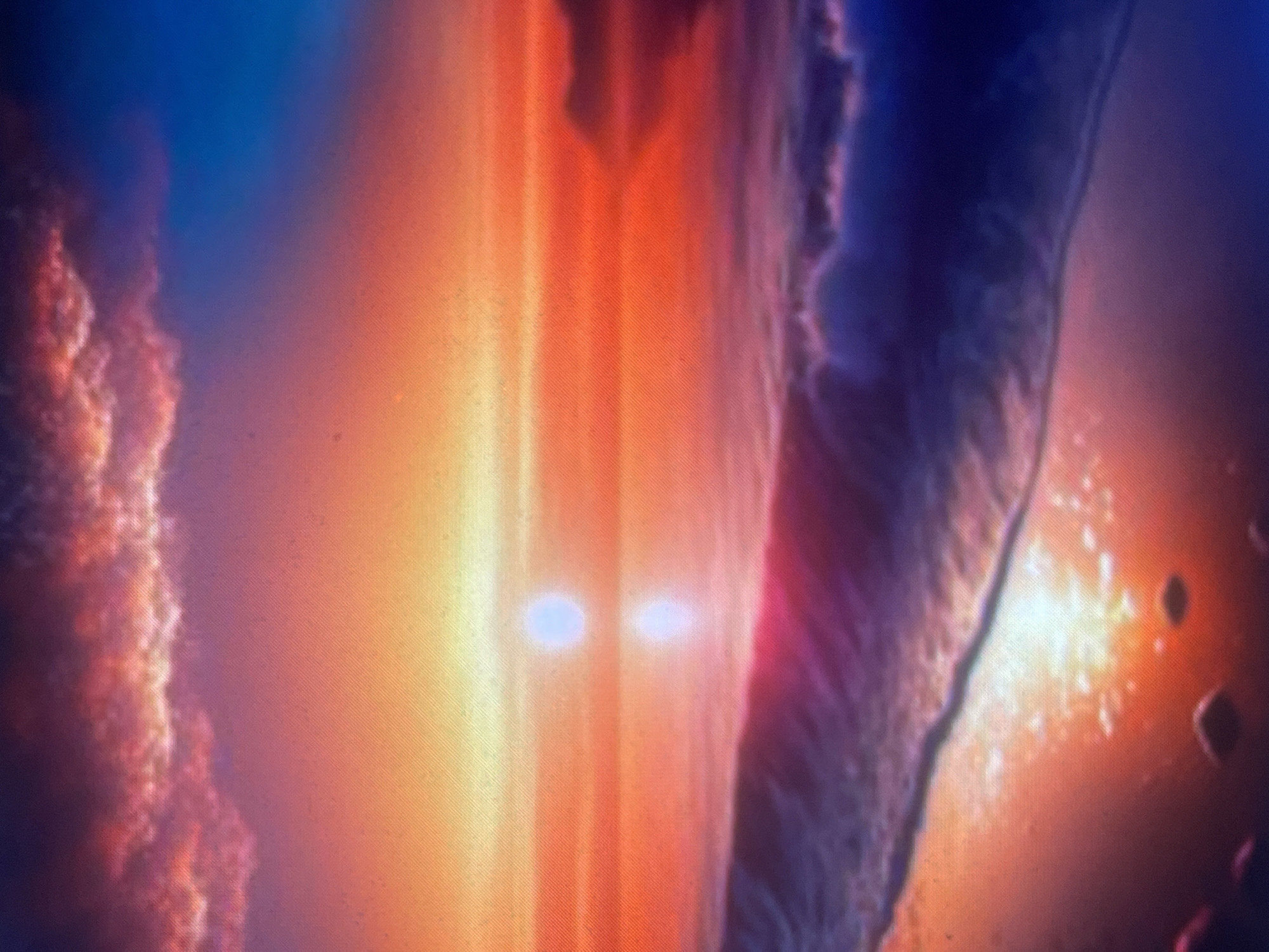 The image depicts a highly saturated coastal sunset or sunrise scene, which is geographically incompatible with any known inland settlement named Zelenyi Hai in Ukraine.
A prominent, bright, circular light source, resembling the sun, is centered on the horizon, casting an intense, shimmering reflection across the water directly below it. The sky displays a dramatic gradient of colors, transitioning from deep blue in the upper right, through rich purples, vivid reds, oranges, and bright yellows towards the horizon. Dark, textured clouds, exhibiting red and purple hues, are visible in the upper left quadrant of the sky.
The body of water in the mid-ground reflects the vibrant sky colors. In the foreground, a substantial wave, characterized by dark purple-blue water and a crest of white foam, is breaking and receding towards the left. The immediate foreground shows a wet, highly reflective sandy beach, mirroring the intense light from the sky and the sun. Several small, dark, irregular shapes, consistent with pebbles or small rocks, are scattered on the wet sand. No people, animals, or man-made structures are visible within the scene.
