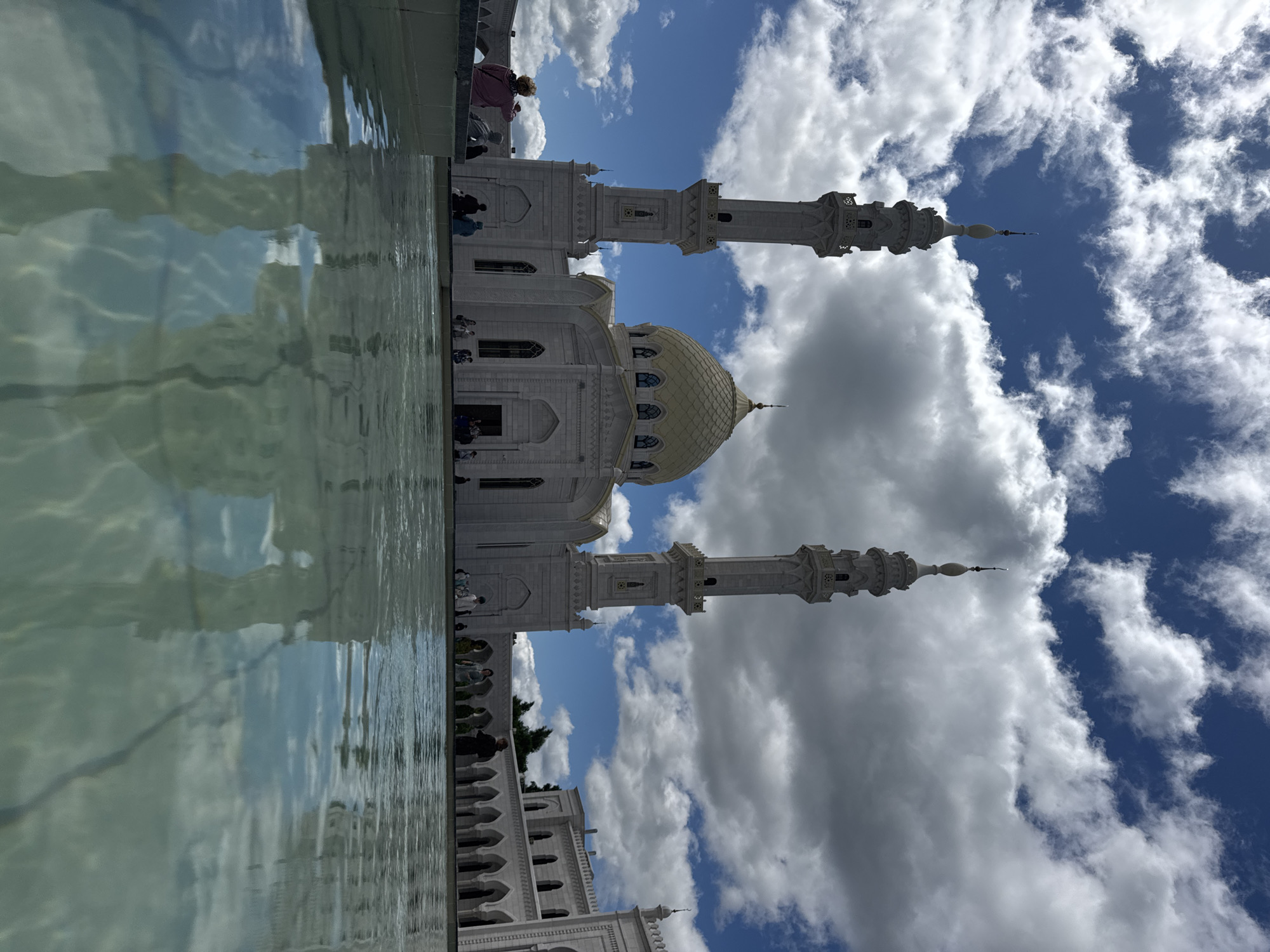 A low-angle, wide shot captures a majestic white mosque with a golden dome and two prominent minarets against a backdrop of a bright blue sky dotted with fluffy white clouds. The mosque, located in the city of Bolgar, Russia, is reflected in the still, clear water of a shallow pool in the foreground. People are visible scattered around the courtyard of the mosque, some sitting by the pool and others walking. The architecture of the mosque is intricate, with arched windows and detailed stonework visible. The overall scene conveys a sense of peace and grandeur.