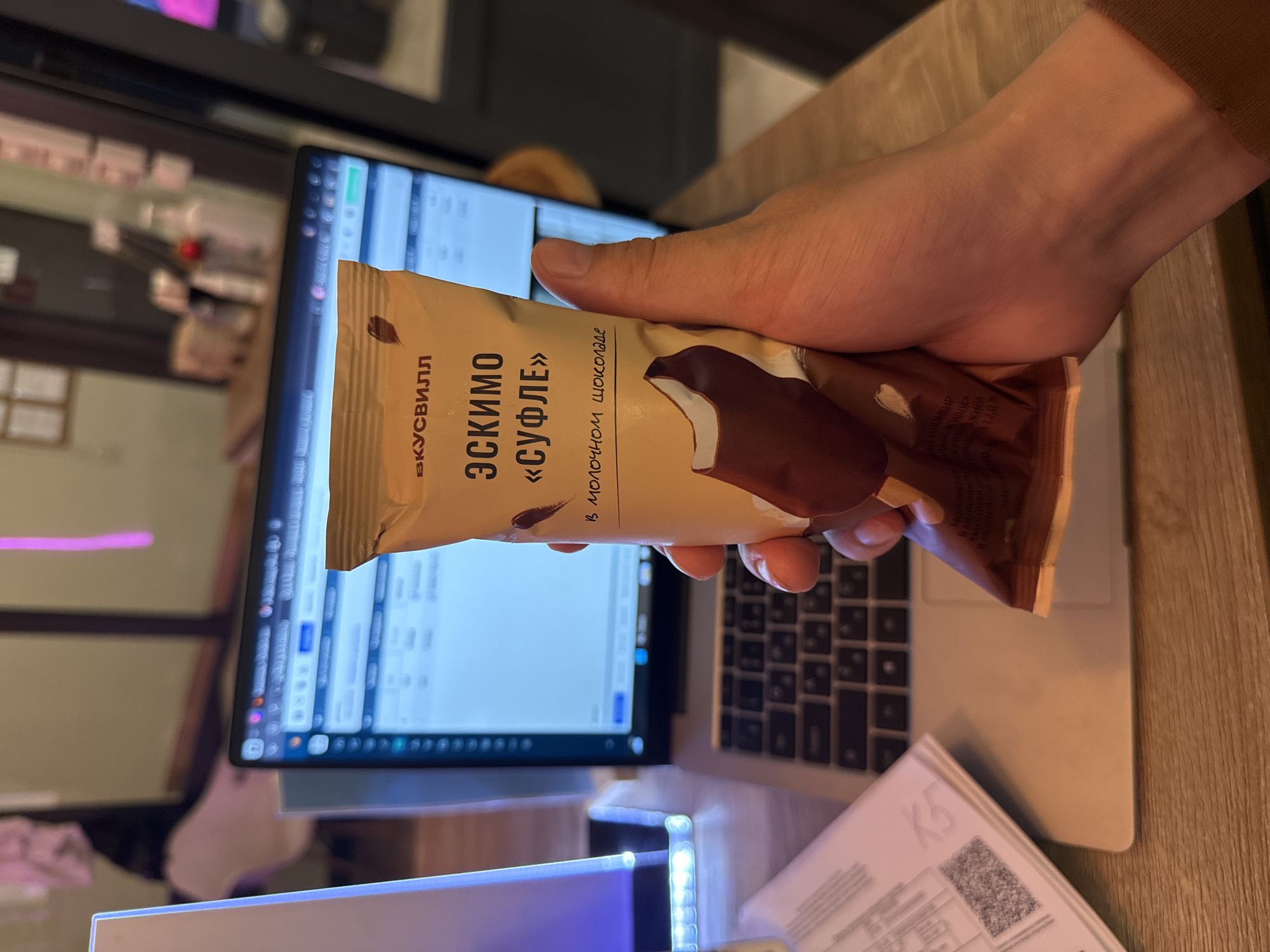 A person's hand is holding a packaged ice cream bar in front of a laptop screen. The ice cream bar has a light brown wrapper with text in Russian. Visible text on the wrapper includes "ВКУСВИЛЛ", "ЭСКИМО", and "«СУФЛЕ»". Below this, there is text that appears to be an ingredient list and nutritional information. The ice cream bar itself is partially visible, showing a chocolate-covered portion.
The laptop screen displays a spreadsheet or document with columns and rows of text and numbers, suggesting the person might be working or studying. The background shows a wooden desk and hints of an indoor environment, possibly an office or home. The lighting suggests it could be daytime, but the specific time or weather cannot be determined. The overall scene appears to be a casual moment of work or leisure, with the ice cream break being a small interruption. The orientation of the image is rotated 90 degrees clockwise.