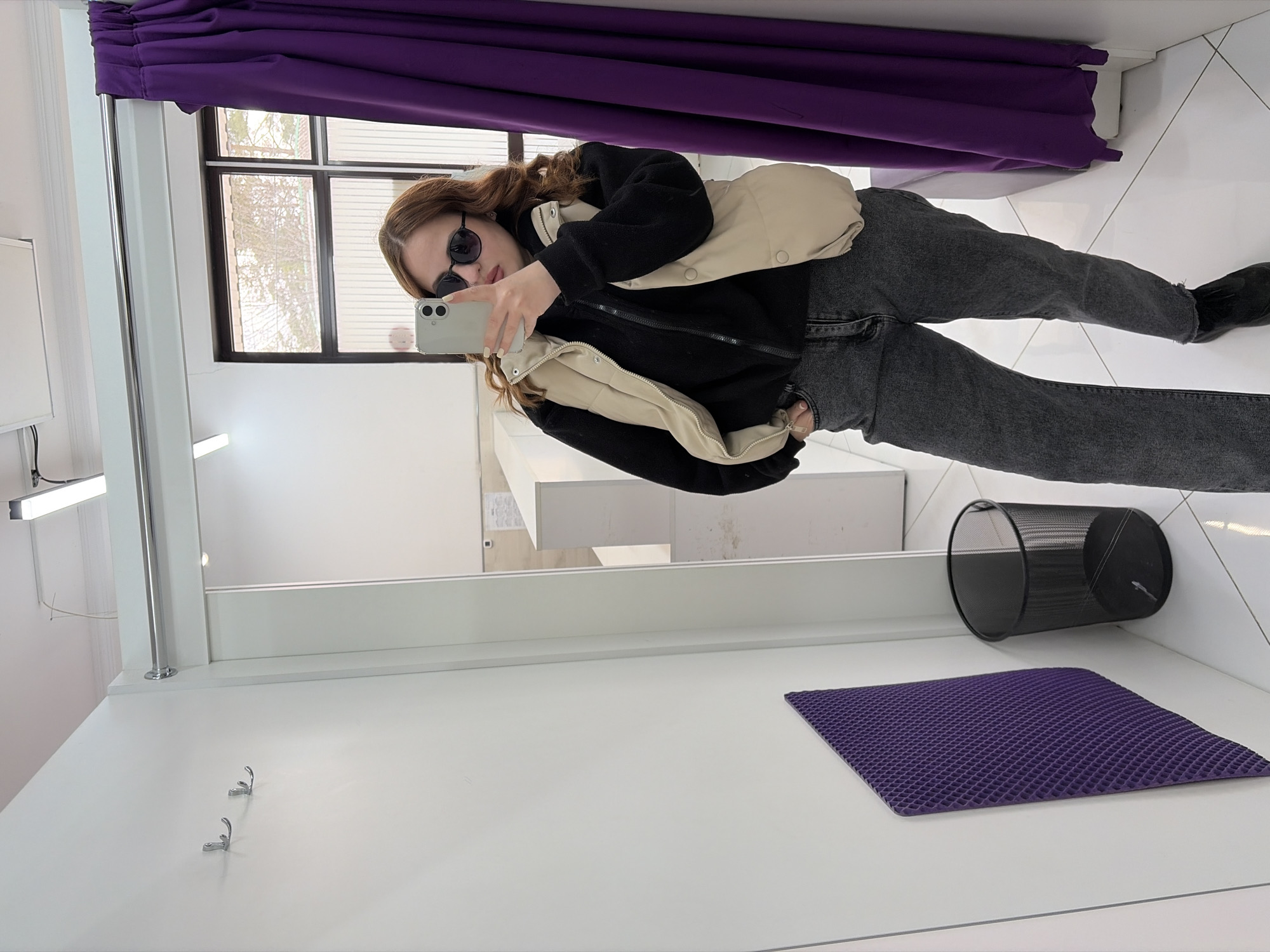 A woman stands in a dressing room, holding a cell phone and taking a photo of herself in a mirror. She is wearing sunglasses, a black jacket, a cream-colored vest, gray jeans, and black shoes. The dressing room features a purple curtain, a mirror, coat hooks on a white wall, a black trash can, and a purple mat on the floor. A window with blinds is visible behind her. The photograph was taken in Novopokrovskaya, Russia.