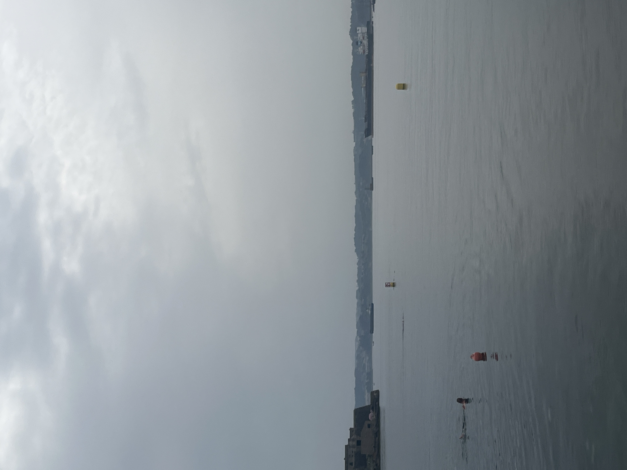 The image depicts a seascape with a hazy, overcast sky. The water is calm and reflects the muted light. On the right side of the frame, a distant coastline is visible, with buildings nestled along a hillside. Several buoys are scattered across the water, some brightly colored, hinting at a designated area or a navigational marker. In the foreground of the water, there are a few buoys that are submerged with only a small portion visible above the water, suggesting they might be part of a buoy line or marker system. One of these buoys is reddish-orange. Further out, a yellow buoy is clearly visible. The overall atmosphere is tranquil and somewhat somber due to the overcast weather. The time of day appears to be daytime, but the lack of direct sunlight and the diffused light suggest it might be morning or late afternoon on a cloudy day. There are no people visible in the image. The scene is likely a harbor or coastal area, given the presence of buoys and the visible coastline with structures. The location context provided is A Coruña, Spain. No text is visible in the image.