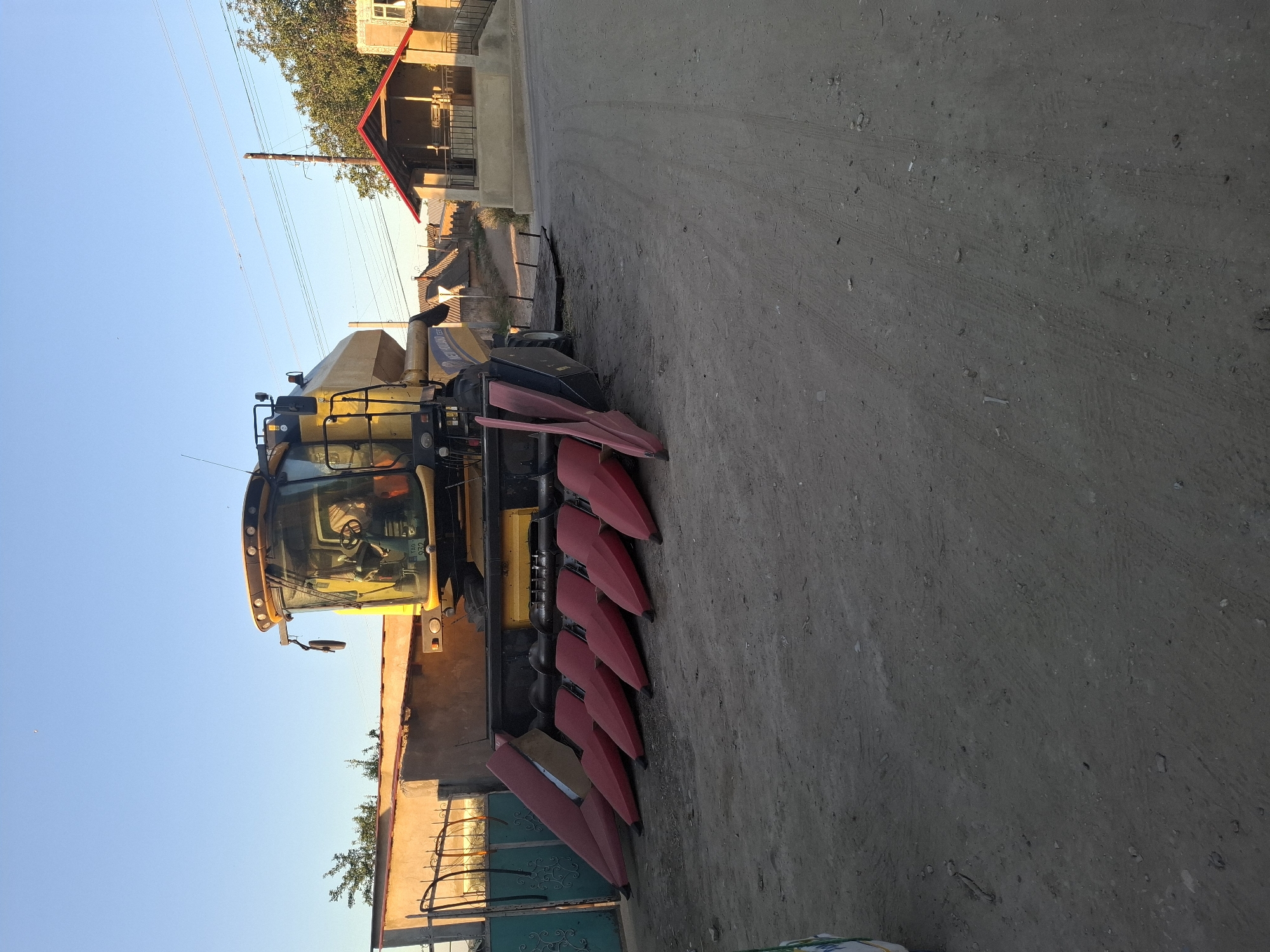 A yellow New Holland combine harvester is parked on a dirt road in Dubăsarii Vechi, Moldova, with a clear blue sky overhead. The combine features a large header with multiple red cutting units. To the left of the combine, there is a green metal fence and a beige building. To the right, a small structure with a red roof and a balcony is visible, along with other houses and trees in the background, suggesting a rural or semi-rural setting. The ground is uneven and appears to be dusty. The scene is illuminated by natural sunlight, indicating it is daytime.