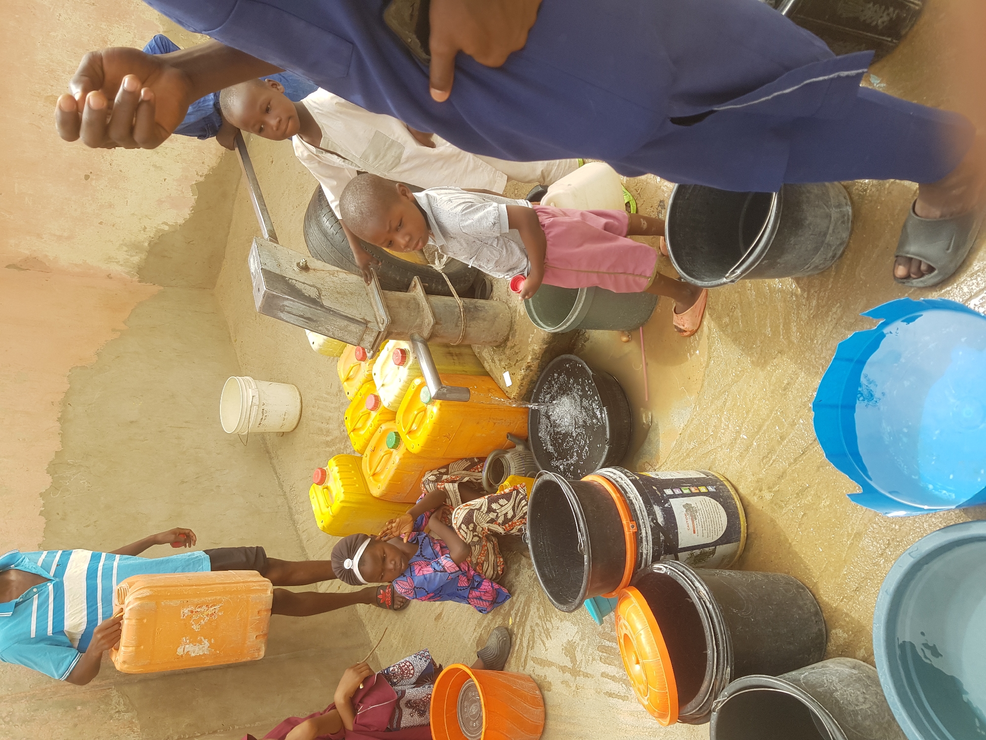 The image captures a scene in Dutsinma B, Nigeria, where several people, including children, are collecting water. A water pump, situated in the center, is dispensing water into various containers: buckets, yellow jerrycans, and other receptacles. The ground is wet, presumably from spilled water. Several individuals, including adults and children, are actively filling containers. Two adults lie on the floor helping with water collection. A child is standing on a tire. Another person is visible on the left holding an orange jerrycan. The setting appears to be an outdoor area, enclosed by a wall. The lighting is bright, indicating daytime.