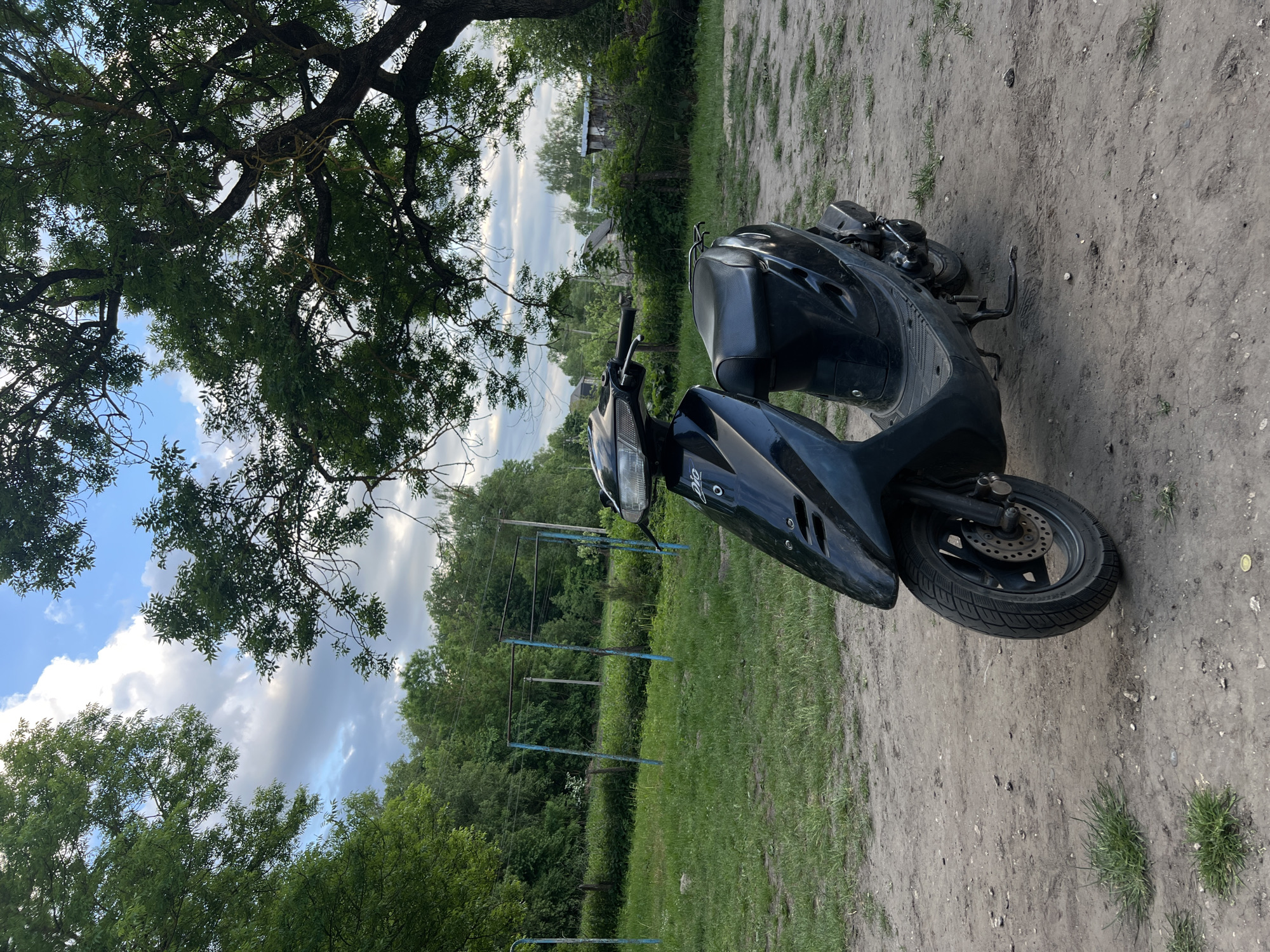The image shows a black moped parked on a dirt and grassy area. A large tree with full foliage is to the left, and a clothesline structure stands in the background. In the distance, there are trees and a building under a cloudy sky.