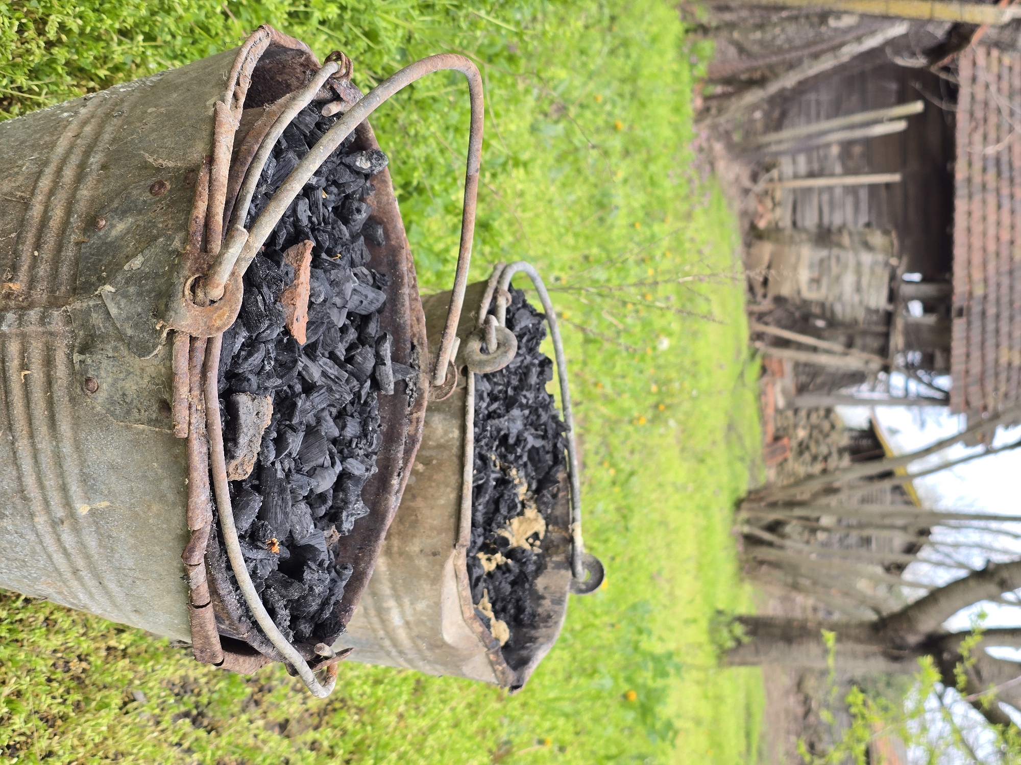 Two galvanized steel buckets sit on green grass outside a dilapidated structure with a tiled roof, possibly in a rural setting. The buckets are filled with what appears to be charcoal or burnt wood. Both buckets show signs of rust, and the handles are visibly corroded. The structure in the background is wooden, with some boards missing, revealing an interior that is partially obscured. Firewood is stacked to the side of the building. The setting appears to be in a state of disrepair and neglect. Location: Unknown, Unknown.