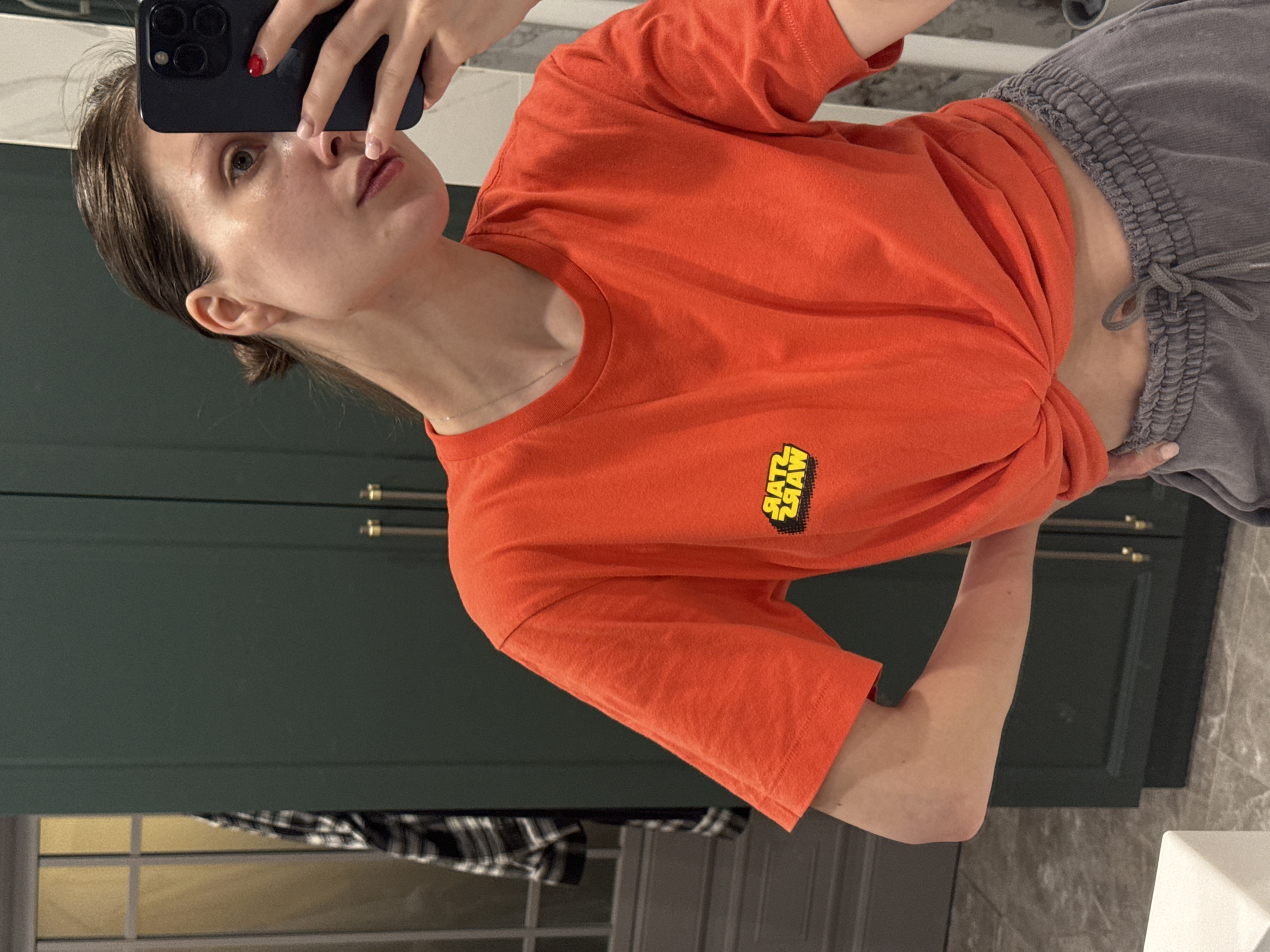 A young adult female is taking a selfie in a bathroom. The background shows a dark green cabinet. The floor and wall surfaces are made of light gray marble. The woman is wearing a red T-shirt with a Star Wars logo on it and gray sweatpants. She is holding a dark-colored smartphone to take the picture. The location is not discernible as Tomilino, Russia based on the visual information provided.