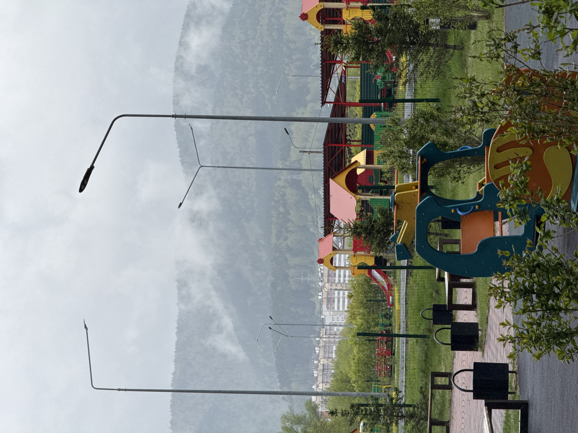 The image shows a playground in Krasnoyarsk, Russia, with a mountain partially obscured by clouds in the background. Several playground structures are visible, including a slide and other colorful play equipment. A row of benches and trash receptacles line a paved path. Streetlights stand adjacent to the playground area. In the background, there are several tall white buildings, likely apartment blocks, and greenery. The sky is overcast.