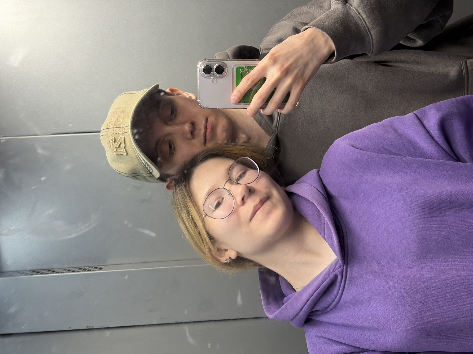 A young couple is taking a selfie in an elevator. The woman is on the left, leaning her head on the man's shoulder. She is wearing a purple hoodie and round glasses. The man is on the right, wearing a grey hoodie and a baseball cap. He is holding a phone to take the picture. The elevator walls are made of brushed metal.