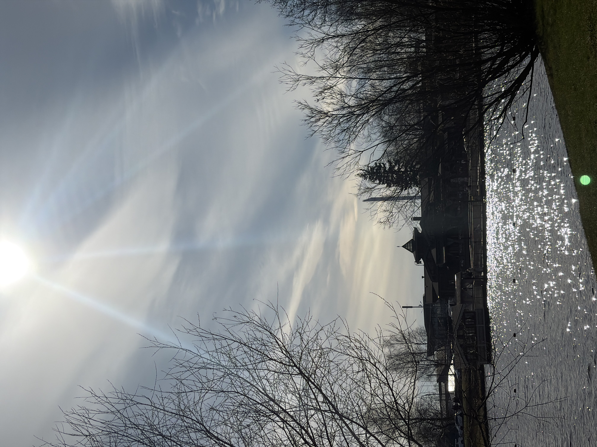 The image depicts a waterfront scene in St. Petersburg, Russia, on a bright day. The sun is low in the sky, shining through the clouds and creating a glare on the water surface. Bare trees line the waterfront. A building with a distinctive tower can be seen on the waterfront. The sky is a pale grey-blue with wisps of clouds. The water is dark grey, with bright highlights from the sun.