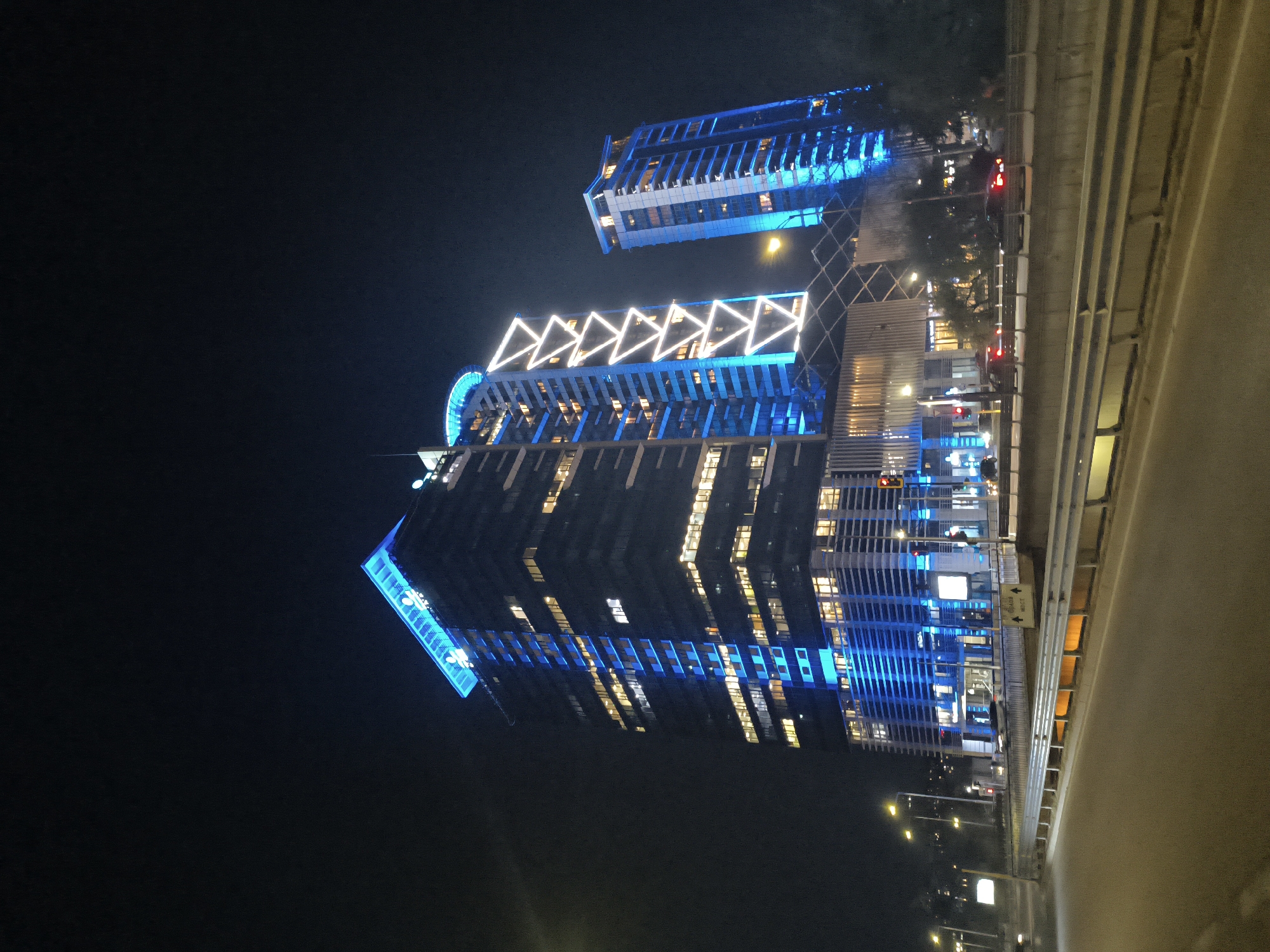 The image shows two tall buildings illuminated with blue and white lights at night. They appear to be modern architectural structures with a combination of lit windows and decorative lighting. A road or highway is visible in the foreground, possibly a bridge or overpass. Streetlights and traffic signals are present, indicating a city environment. Based on the architectural style and lighting, the buildings are likely located in Sofia, Bulgaria. The illumination suggests a planned light show or festive decoration.