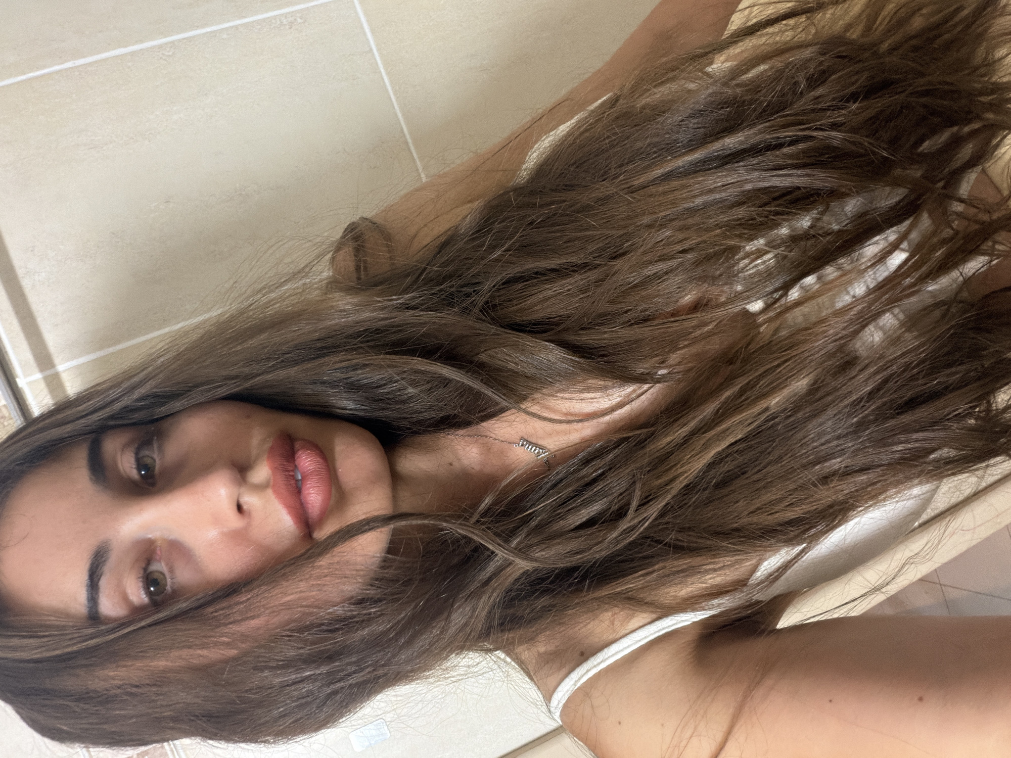 The media file is a close-up, overhead shot of a young woman lying down. She has long, dark brown hair that is partially obscuring her face and body. Her face is turned towards the camera, and she has green eyes, brown eyebrows, and is wearing pink lipstick. She is also wearing a white tank top and a delicate silver necklace with small charms. The background appears to be a tiled floor, possibly in a bathroom or kitchen, with beige tiles and white grout lines. The lighting suggests it might be daytime. The woman's expression is neutral or slightly relaxed. There is no visible text in the image. The scene is a personal, candid moment.