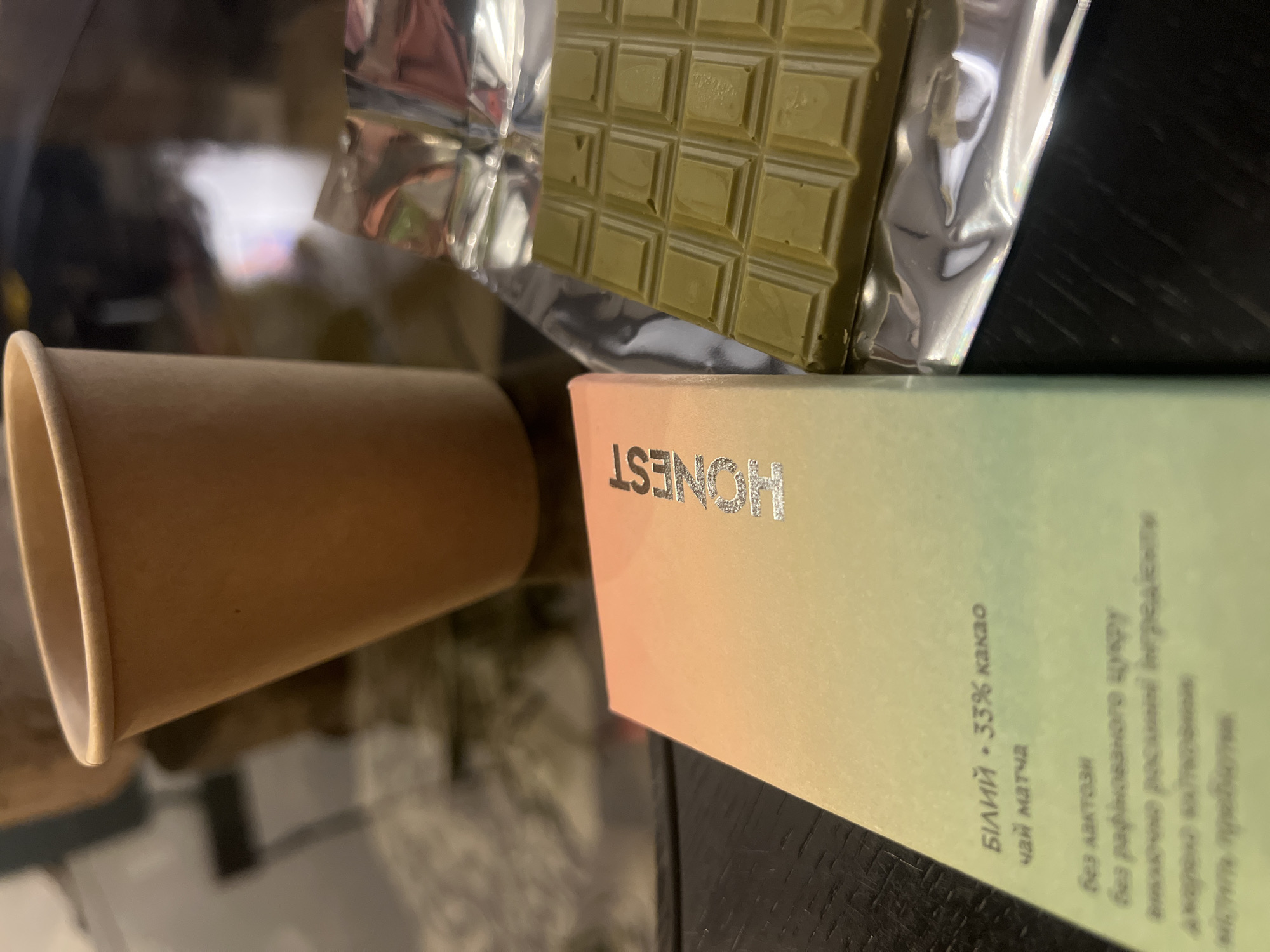 In Kyiv, Ukraine, on a dark, reflective surface, a light brown paper cup stands to the left of a partially unwrapped green chocolate bar. The chocolate bar is segmented into a grid of squares and rests on a silver foil wrapper. To the left of the chocolate bar and partially obscuring the view of the cup is a rectangular cardboard box with a gradient of colors ranging from peach to green. Silver lettering on the box spells out "HONEST". Text in a smaller font is visible on the lower portion of the box, with some of it being illegible due to the focus and angle. The scene suggests a casual setting, possibly a desk or table.