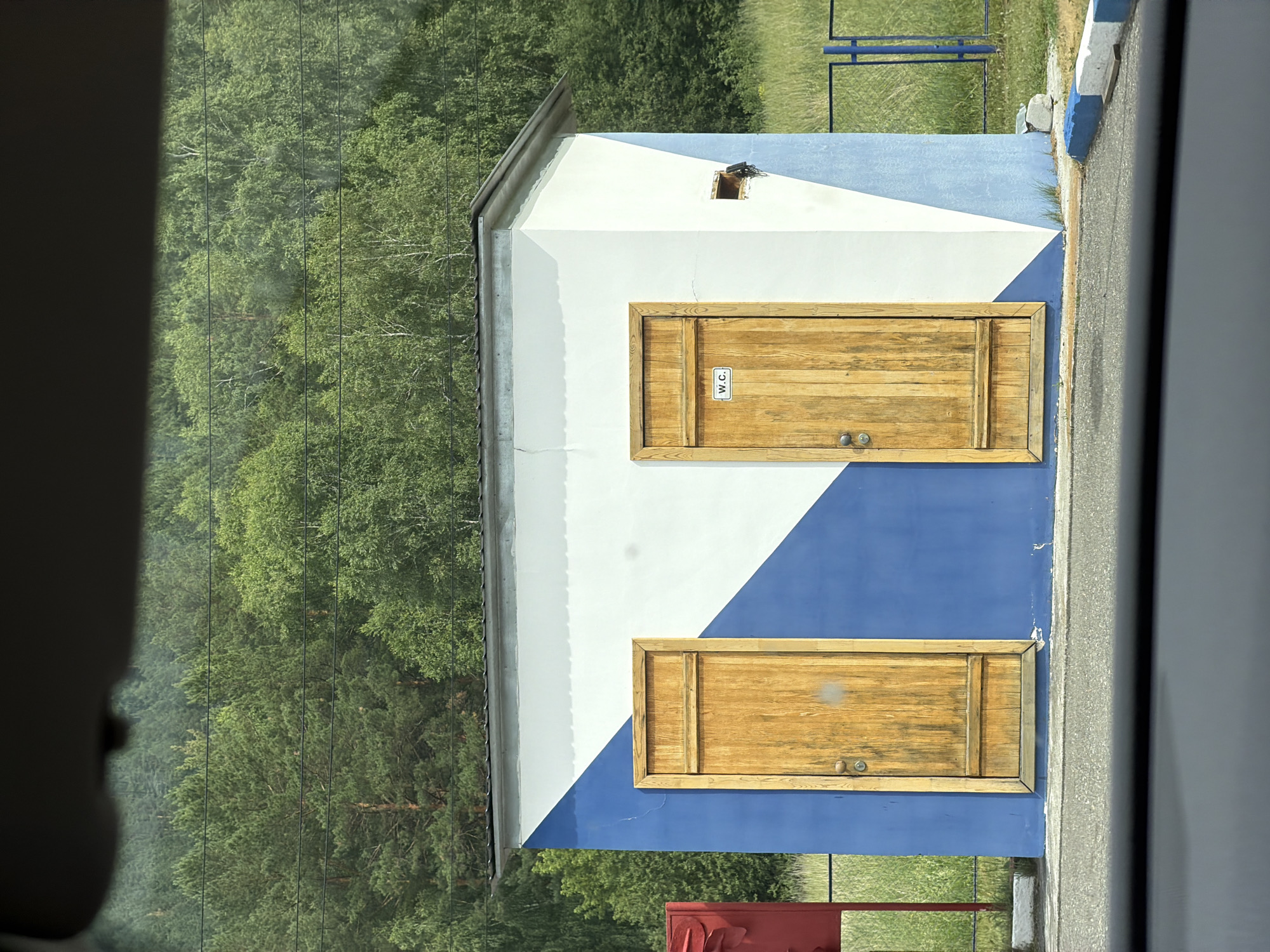 The image shows a small, blue and white building with two wooden doors. The building is located outdoors, in a grassy area with a forest in the background. The building has a pitched roof and a vent on the side. One of the doors has a "W.C." sign on it. A red object and a metal fence are visible to the left of the building. The image appears to have been taken from inside a vehicle, as suggested by the dark shadow at the bottom of the frame and a slight reflection on the surface. The location is identified as Katun, Russia.