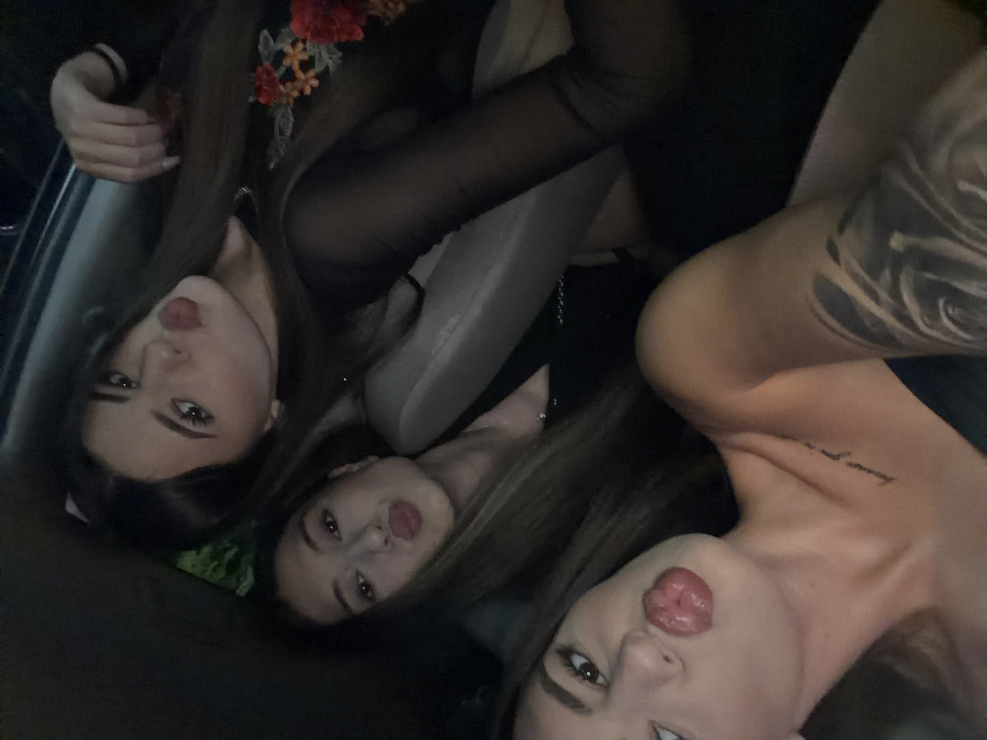 Three young women are seated in the back of a car at night. The woman on the left is closest to the camera, with her head tilted back and her tongue sticking out. She has a tattoo on her arm and collarbone. The woman in the middle is looking at the camera with her lips pursed. The woman on the right is also looking at the camera with her lips pursed, and she is wearing a sheer black top with floral embroidery. The car appears to be parked, possibly in a residential area, given the faint greenery visible in the background. There is no specific indication of the location being Plovdiv, Bulgaria.