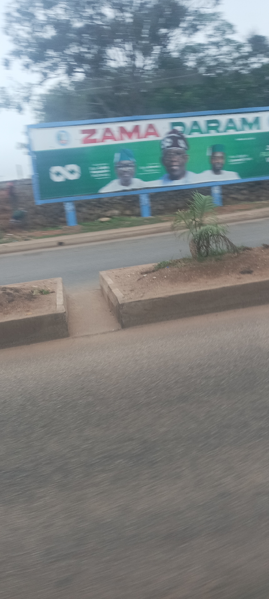 This image captures a roadside scene in Jos, Nigeria, likely taken from a moving vehicle due to the motion blur evident on the asphalt road in the foreground and the heavily blurred trees in the background.
The central subject is a large, green and blue billboard, supported by blue poles, positioned to the right of the road. On the billboard, three male figures are prominently displayed, suggesting they are public or political figures. The central figure wears a patterned cap, while the man to his left wears a light blue cap, and the man to his right wears a green cap. The most prominent text on the billboard, in red and white letters, reads "ZAMA DARAM". A white symbol resembling an infinity sign or the number '8' is also visible on the left side of the billboard, alongside a logo at the top left.
Between the road and the billboard is a concrete median or curb, with a patch of dirt where a small, palm-like plant is growing. In the far distance, blurred foliage indicates trees under an overcast or hazy sky, contributing to the diffused lighting of the scene. A very indistinct, dark human figure can be faintly seen behind the left end of the billboard. The scene depicts a typical street environment featuring public messaging.