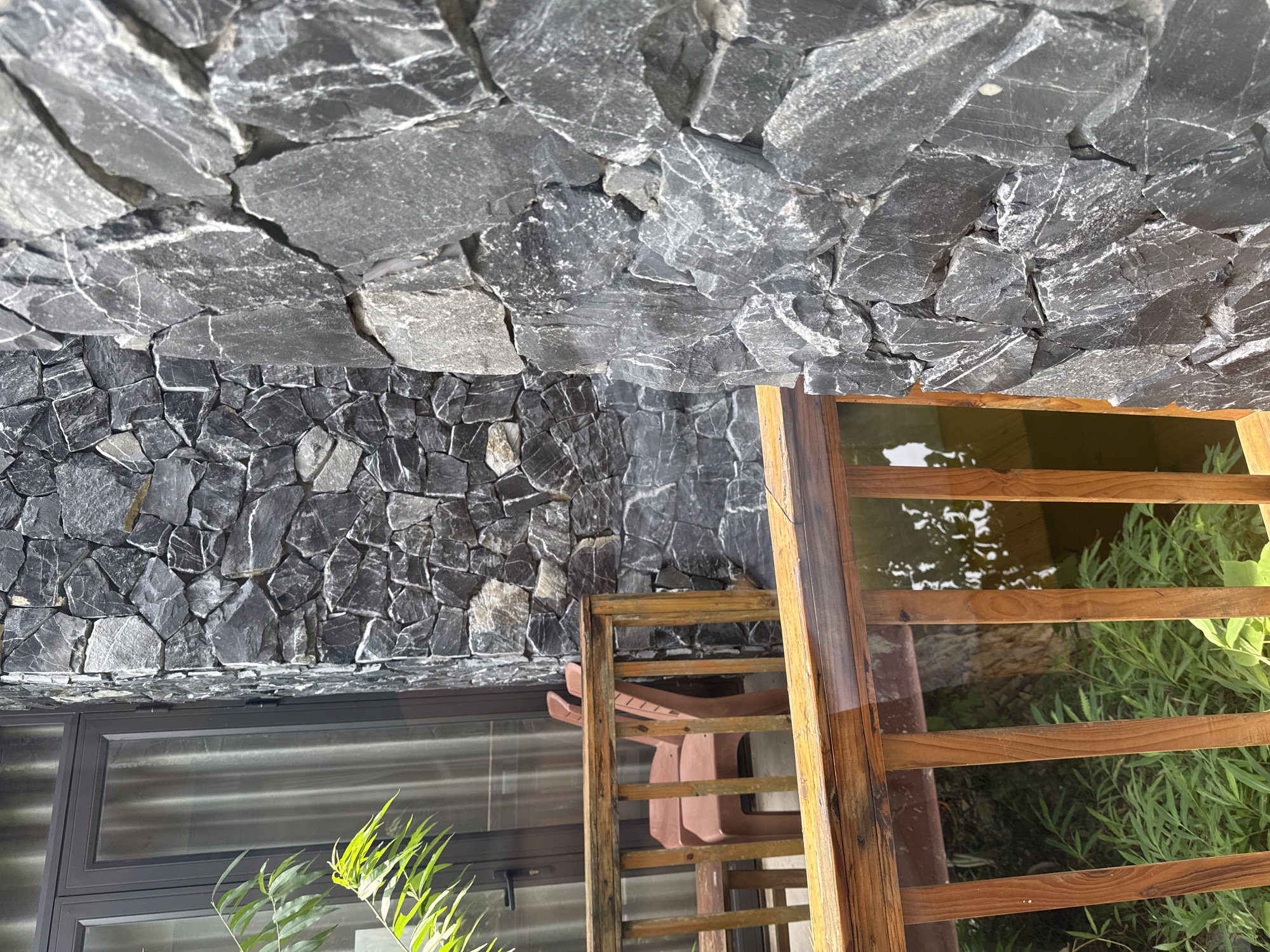 The image displays an outdoor scene featuring a stone wall, a wooden fence, and natural foliage under bright daylight. The dominant feature is a tall wall, constructed from irregular, dark grey and black natural stones. The upper part of the wall consists of larger, flatter stones with noticeable white veins and streaks, while the lower section transitions to smaller, more jagged stones.
To the right, a warm brown wooden fence or railing, made of horizontal slats, runs vertically through the frame. Behind this wooden structure lies a calm body of water, possibly a pond or canal, reflecting light and containing some green aquatic plants. Lush green foliage, resembling tall grass or reeds, grows along the water's edge.
On the left side of the image, a modern window with dark grey frames is visible, hinting at an adjacent building. In the immediate foreground on the left, green leafy plants are present. A reddish-brown outdoor chair is partially visible behind the wooden railing.
The scene is illuminated by natural sunlight, suggesting a clear and sunny day, likely in the late morning or afternoon. There are no individuals or discernible activities taking place. No text is visible. The setting, identified as Du Già Commune, Vietnam, combines natural building materials like stone and wood with contemporary architectural elements.