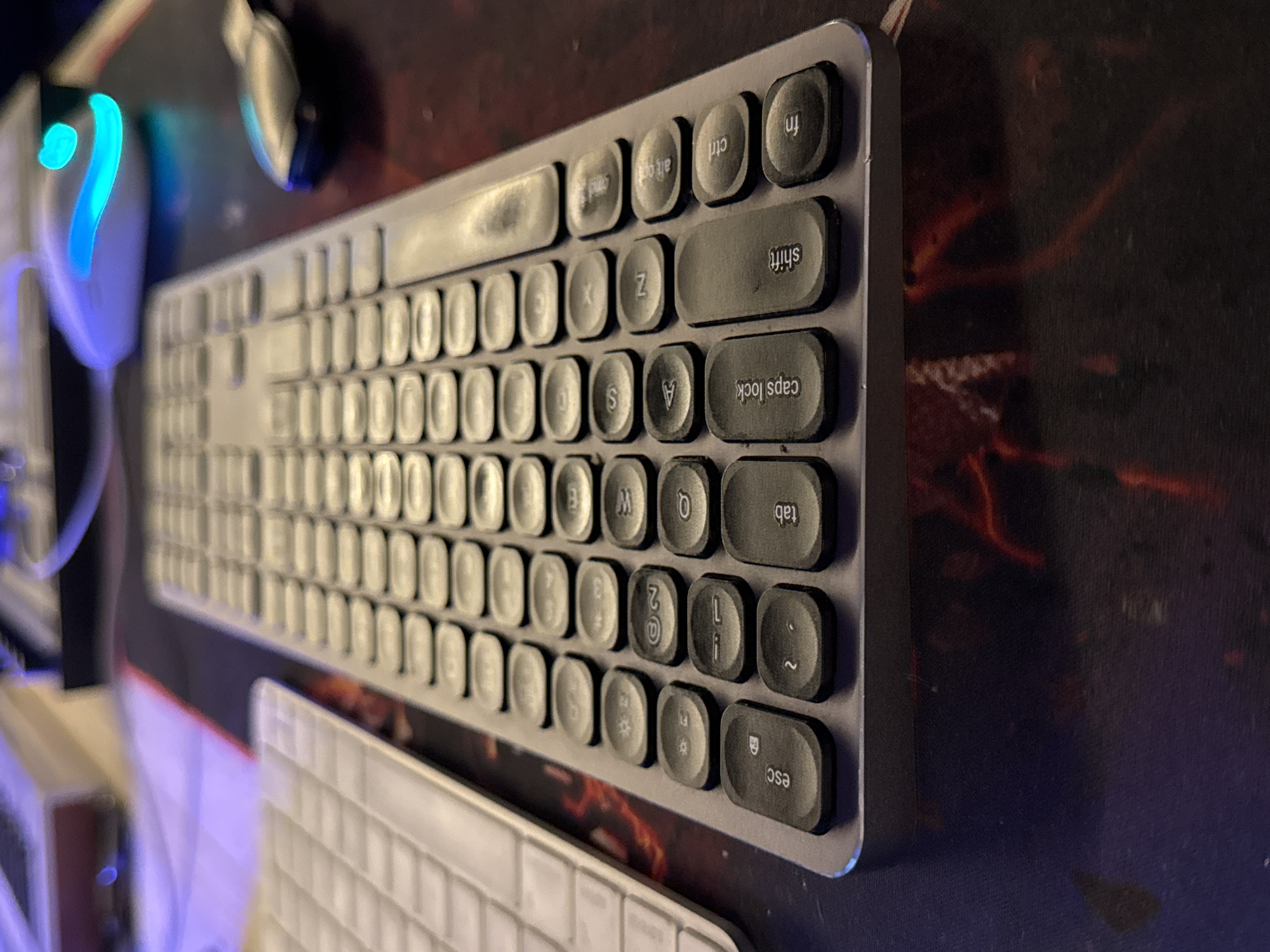 This media file is an eye-level, close-up shot of a computer keyboard and a computer mouse. The keyboard is silver with round, black keys. The keys visible include "fn", "ctrl", "alt gr", "alt", "shift", "caps lock", "tab", and "esc". The mouse is white with blue and green light emanating from it. The keyboard and mouse are resting on a dark patterned desk mat. In the background, a portion of another white keyboard is visible. The overall scene suggests a gaming or work setup. The lighting is dim with blue and green hues from the mouse. The image appears to be taken indoors. No people are visible. The location context is Plovdiv, Bulgaria.