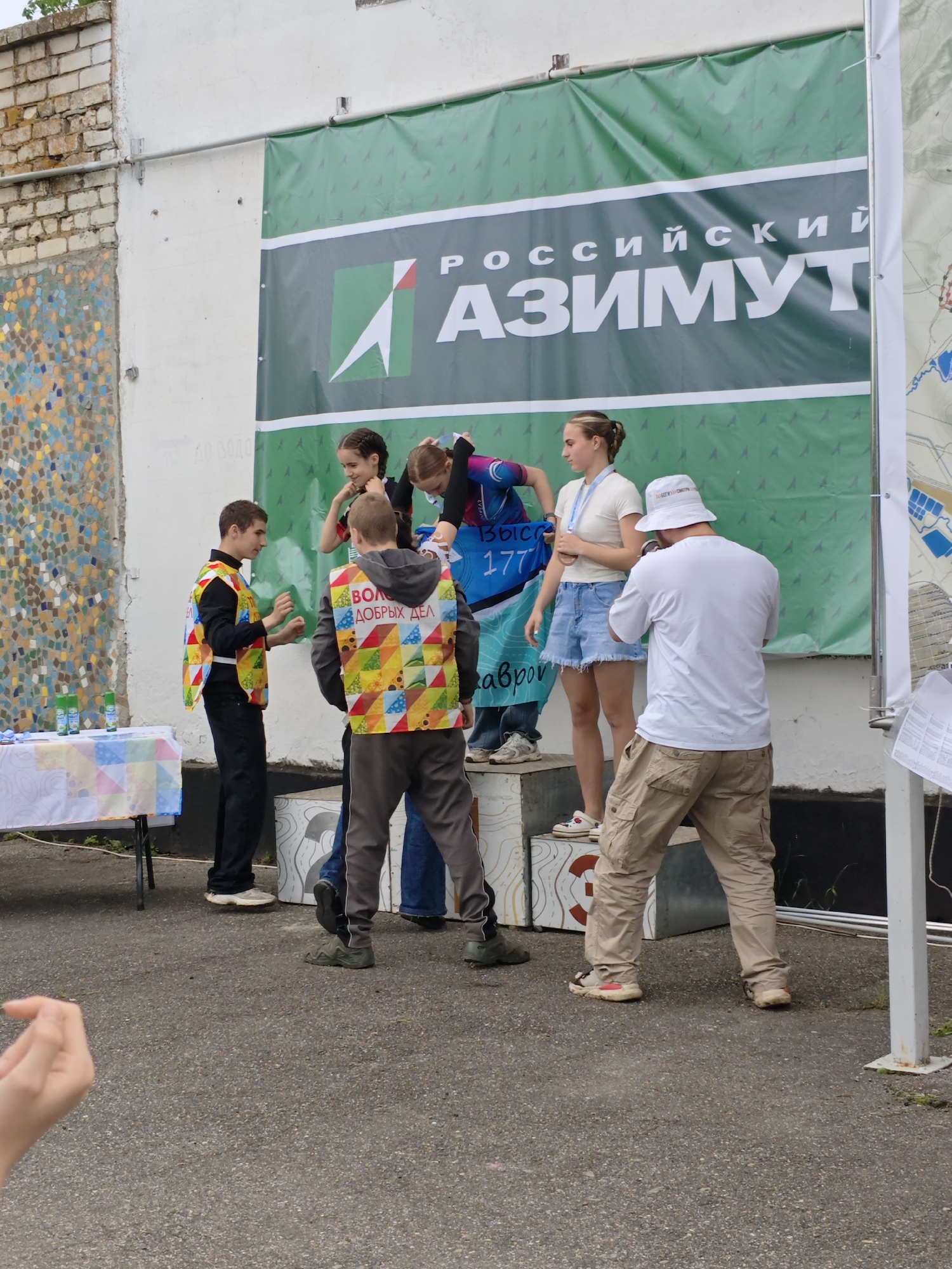 The image shows a group of people standing on and around a platform, possibly a podium for an event. A banner with Russian text, "РОССИЙСКИЙ АЗИМУТ" (RUSSIAN AZIMUT), is behind them. Three young women are on the podium, one wearing a cycling outfit and another with a medal around her neck, suggesting a competition or award ceremony. Two men wearing vests with colorful geometric patterns and the words "ВОЛОНТЕР ДОБРЫХ ДЕЛ" are standing next to the podium, interacting with the people on the platform. Another man in a white t-shirt and bucket hat is facing away from the camera, near the edge of the podium.
To the left of the scene, there is a table with drinks. The background includes a brick wall with a mosaic design and a map. The location can be determined as the city of Новоекатериновская, Russia, based on the query.