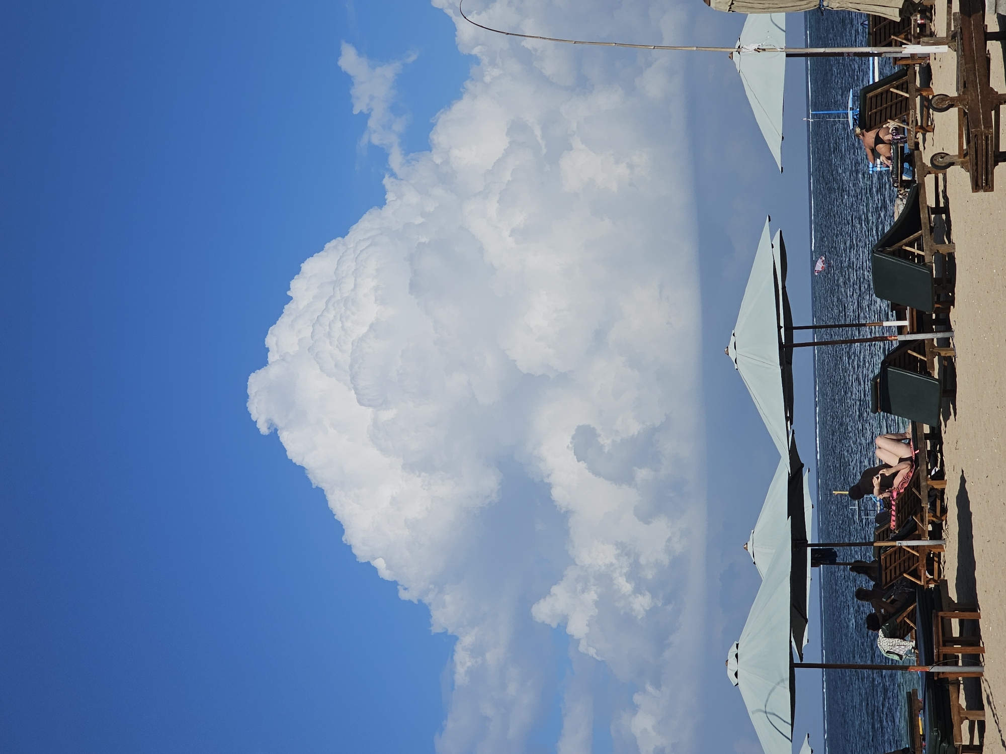 The image depicts a beach scene in Denpasar, Indonesia, under a vibrant blue sky with a massive cumulus cloud formation dominating the upper portion. In the foreground, a sandy beach is lined with several empty and occupied lounge chairs and umbrellas, suggesting a resort or public beach area. The ocean, appearing dark blue with white caps, stretches to the horizon. Several people are visible relaxing on lounge chairs under the shade of the umbrellas. One person is lying on a lounge chair, seemingly reading or looking at their phone, while another is adjusting their attire. A thin, bamboo-like pole extends from the right side of the frame upwards. The overall atmosphere is one of a tranquil beach day.