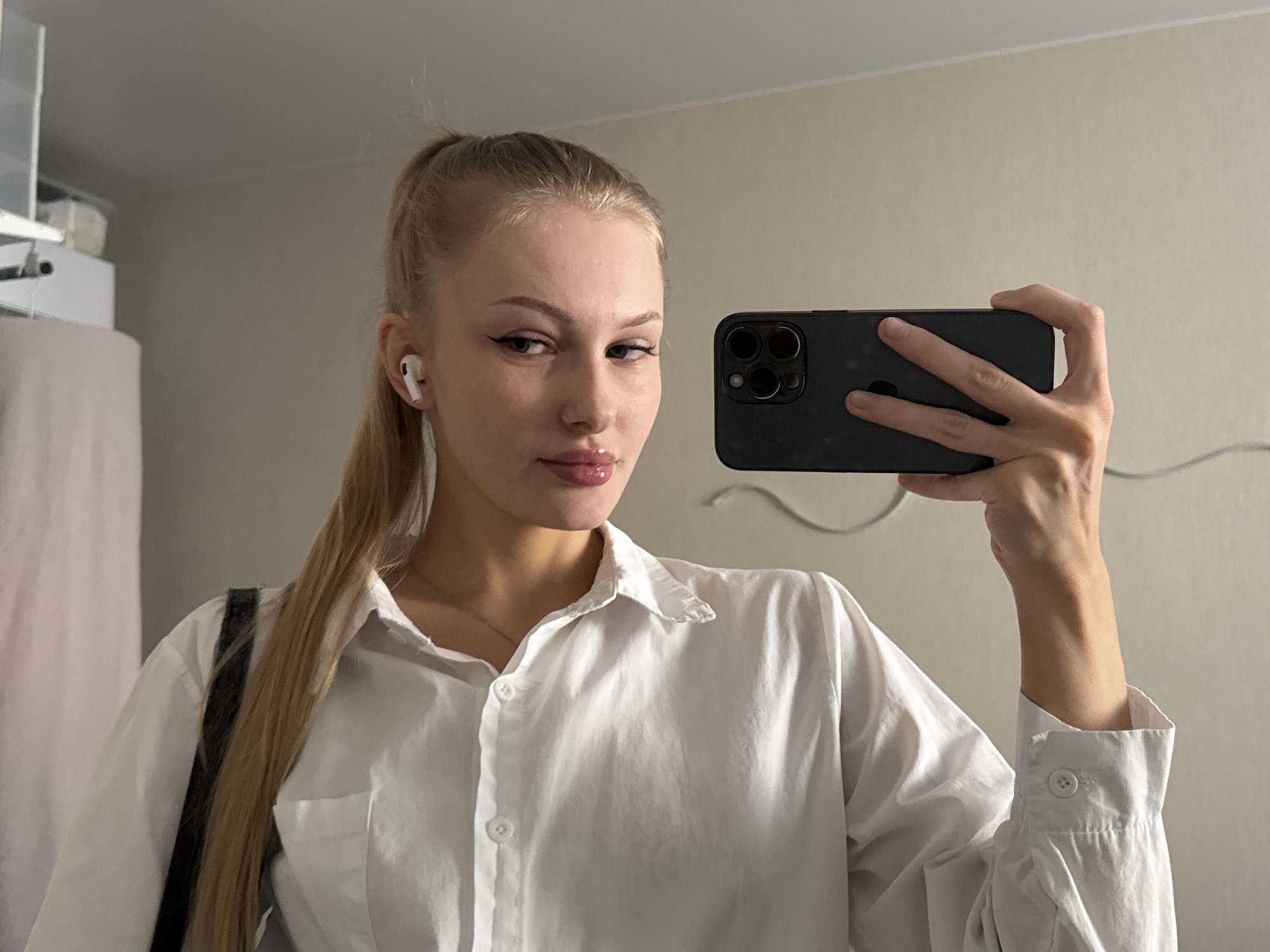 This is a mirror selfie taken indoors, likely in a residential setting. The subject is a young Caucasian woman with blonde hair tied back in a high ponytail. She is wearing a white collared shirt and appears to be wearing white wireless earbuds. She is holding a dark-colored smartphone in her right hand, using it to take the photo. Her makeup includes winged eyeliner and lip gloss. The background is a plain, neutral-colored wall, with a hint of a light-colored curtain or fabric to the left of the frame. There are no specific indications of the time of day or weather. The overall impression is that of a casual, everyday moment.