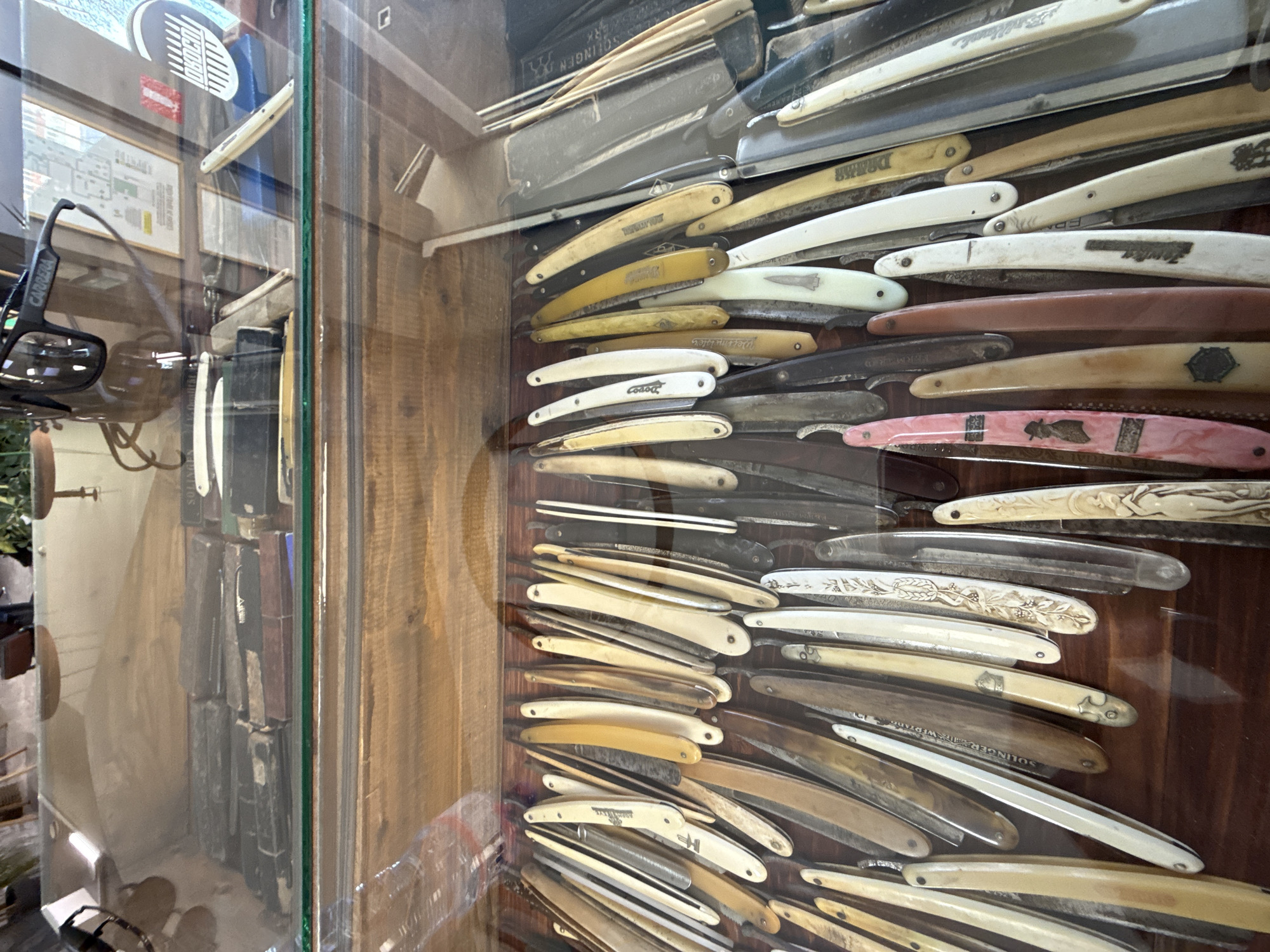 The image shows a display case filled with straight razors of varying styles and ages. The display case is likely in a shop or museum. The reflection in the glass suggests the scene is in Veliko Tarnovo, Bulgaria, as there is an image of a map of the city and a sign that appears to say "Local." The background visible through the glass includes trees. The focus of the image is on the large collection of razors within the case. The razors have different colored handles and some appear to have ornate designs.