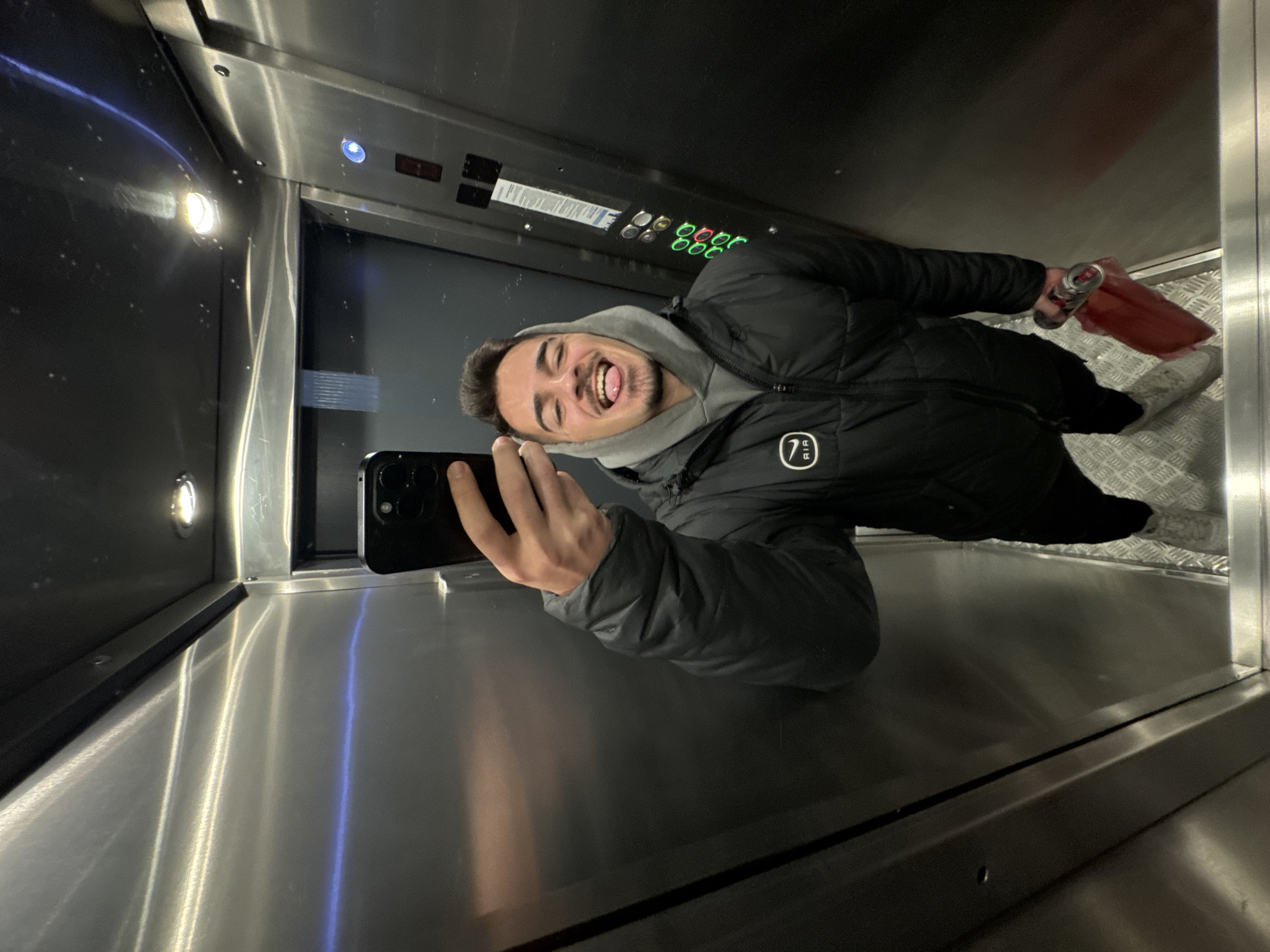 A young man is taking a selfie in an elevator. He is wearing a black puffer jacket over a grey hoodie. He is sticking his tongue out. He is holding a black smartphone in his left hand. In his right hand, he holds a can of what appears to be an energy drink inside of a red carrier bag. The interior of the elevator appears to be metal. Elevator button panel can be seen. The elevator floor has a metallic, diamond-patterned texture. No location information can be determined.