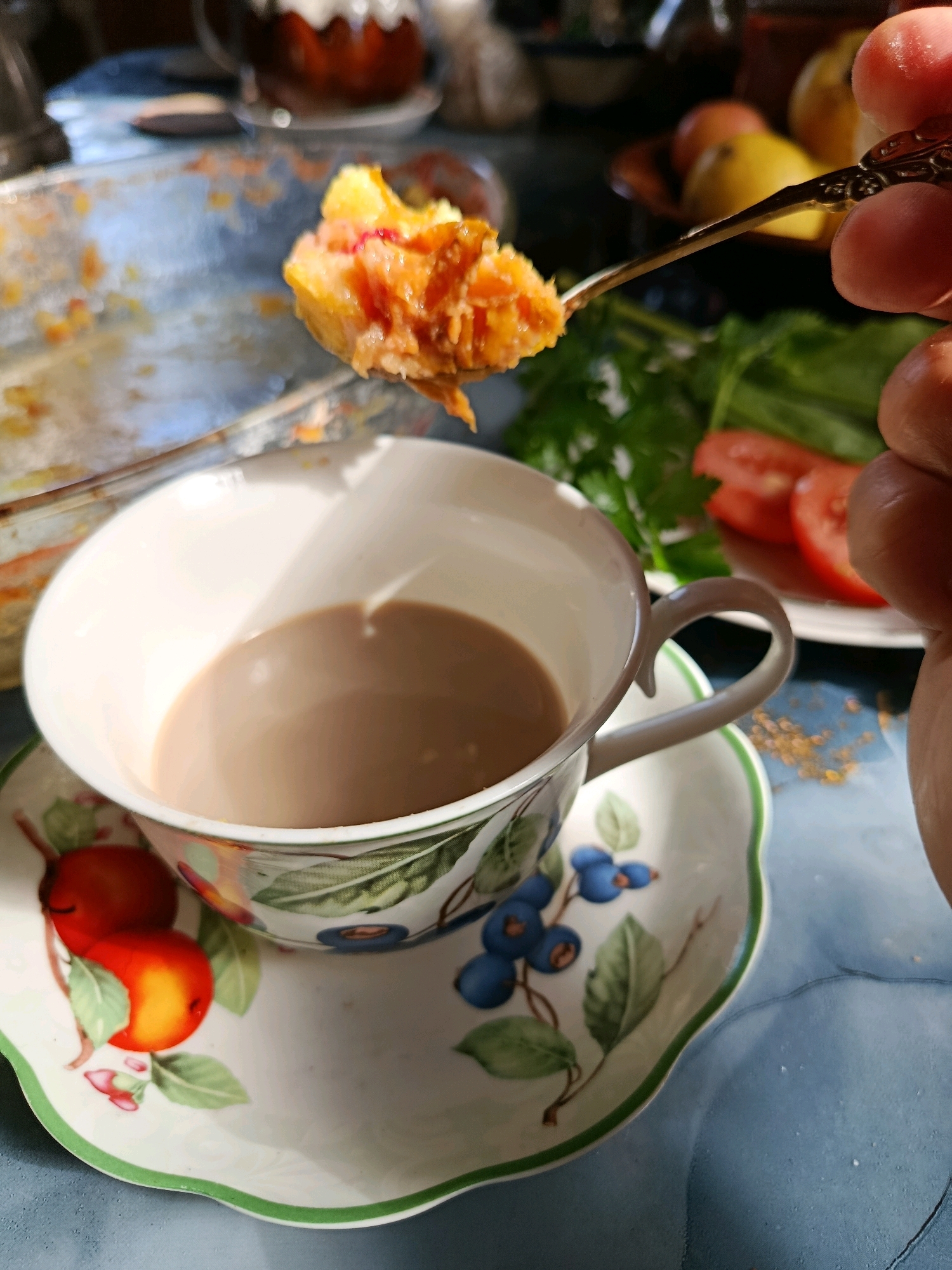A close-up shot captures a forkful of crumbled orange dessert being held over a white teacup filled with light brown liquid. The teacup and saucer feature a floral and fruit pattern with red apples and blueberries. In the background, partially visible, are sliced tomatoes, green leafy vegetables, and a blurry glass object with a brown interior. The scene is set on a blue, slightly textured surface, possibly a table, with sunlight casting shadows. The overall impression is a domestic setting, likely a meal or snack. There is no identifiable information about the location in Vareya, Russia.