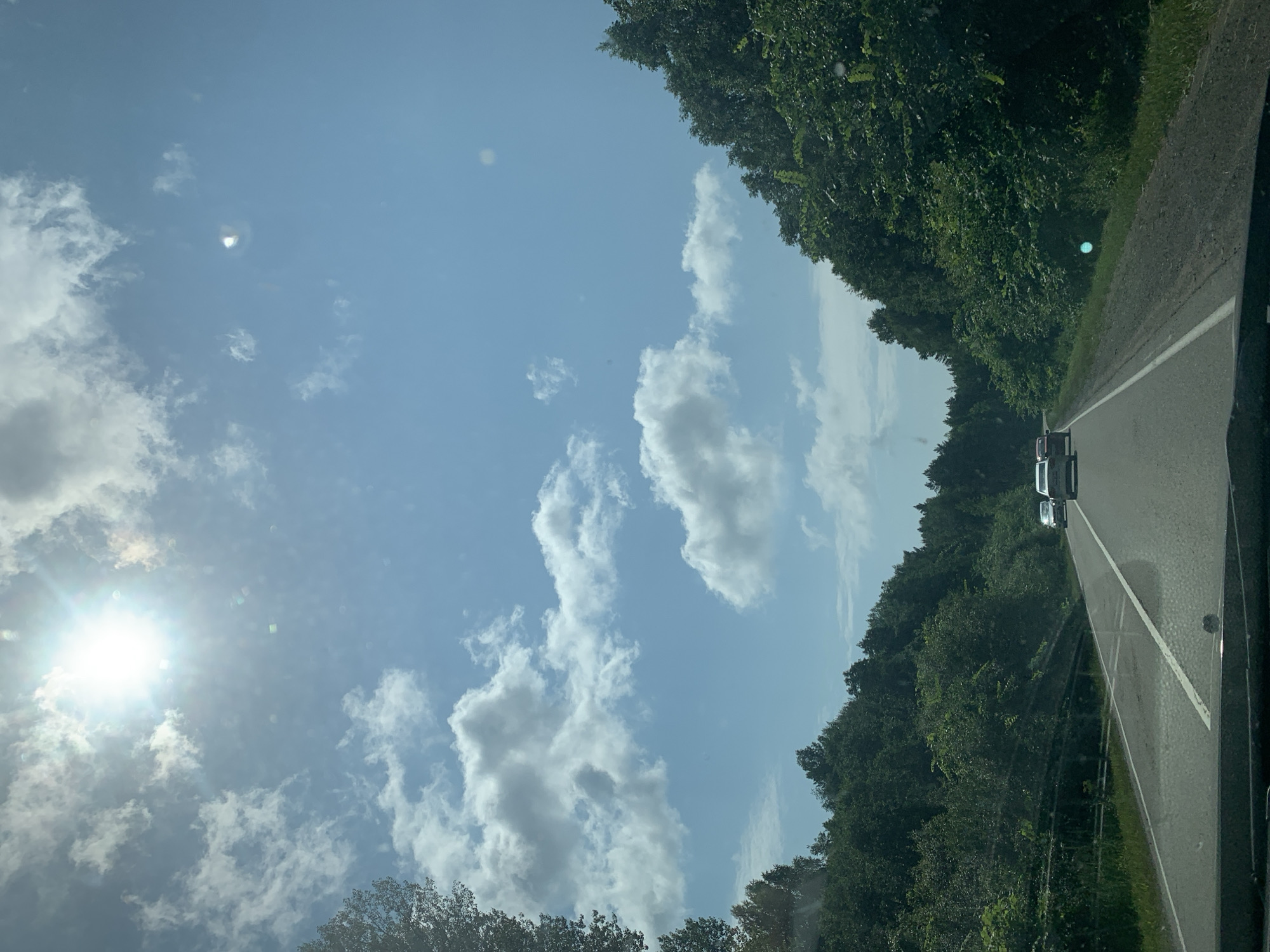 The image depicts a view from inside a vehicle traveling on a highway. The road is asphalt, with white lane markings and a guardrail visible on the left side. Dense green trees line both sides of the road, forming a canopy that extends into the distance. The sky is blue with scattered white clouds, and the sun is prominently visible in the upper left quadrant, casting a bright light. Several cars are visible on the road ahead, traveling in the same direction. The scene appears to be a rural or semi-rural setting in Russia, potentially along the Doma 78 Km highway, given the provided context.