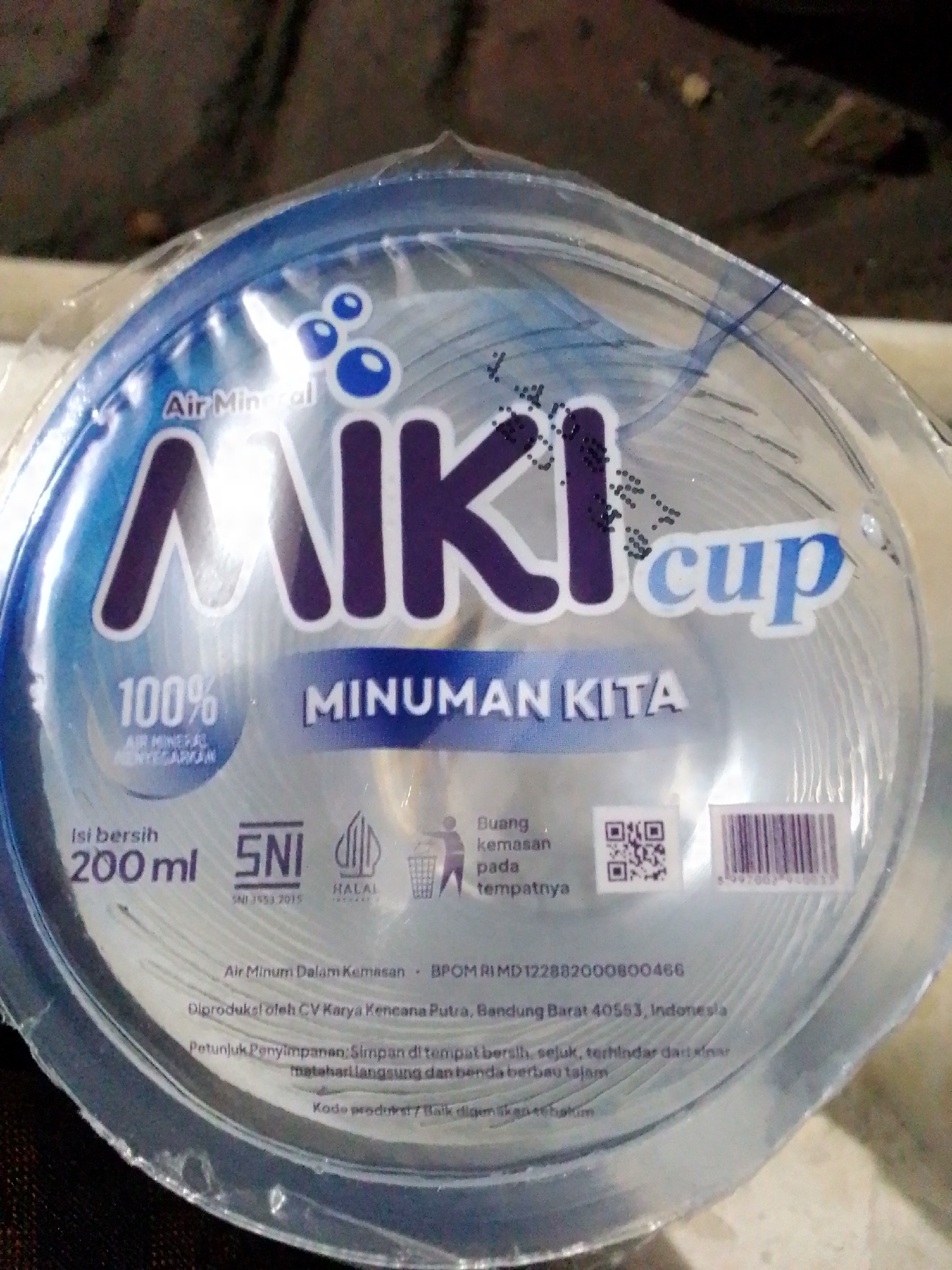 The image shows a close-up of a plastic cup lid for "Miki Cup" brand mineral water. The lid is blue and white with the brand name prominently displayed. There are also English and Indonesian text, as well as a QR code and barcode. The lid is transparent and shows the product inside. The background is blurred and appears to be a domestic setting. There is no visible information about the location being Sukamanah, Indonesia, or any specific details about the environment that can be used to confirm its location.