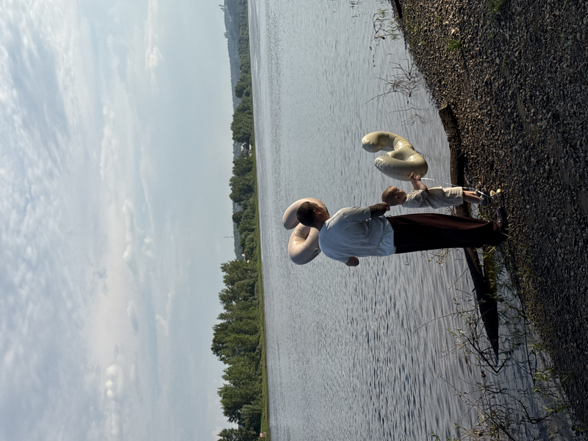 The image captures a man and a young child standing on the rocky shore of the Kama River, near Chaikovsky, Russia. The man, dressed in a light blue t-shirt and dark pants, holds a large, light-colored inflatable ring. The child, wearing a light-colored t-shirt and shorts, is positioned to the man's right and also holds a similar inflatable ring. The child appears to be interacting with the ring, possibly letting water out. Behind them, the Kama River stretches out, with a tree-lined bank visible in the distance, dotted with some structures. The sky is partly cloudy, with a soft, diffused light. The overall scene depicts a casual moment by the water, likely during the daytime.