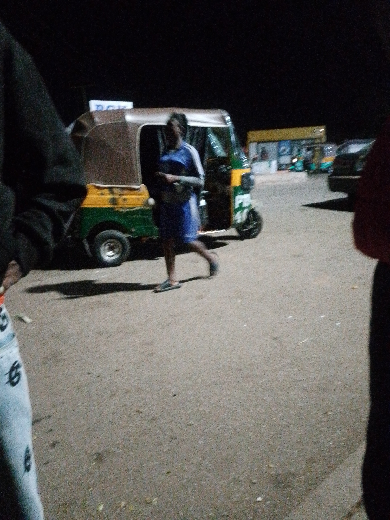 In the city of Unknown, Unknown, an outdoor, ground-level shot captures a nighttime scene. A person, wearing a blue garment and dark footwear, walks across an asphalt surface next to a yellow and green auto-rickshaw. The auto-rickshaw has a brown canopy and its rear wheel is visible. In the background, a building with a yellow overhang and signage can be seen, along with other auto-rickshaws and a dark-colored vehicle. The lighting conditions suggest it is nighttime.