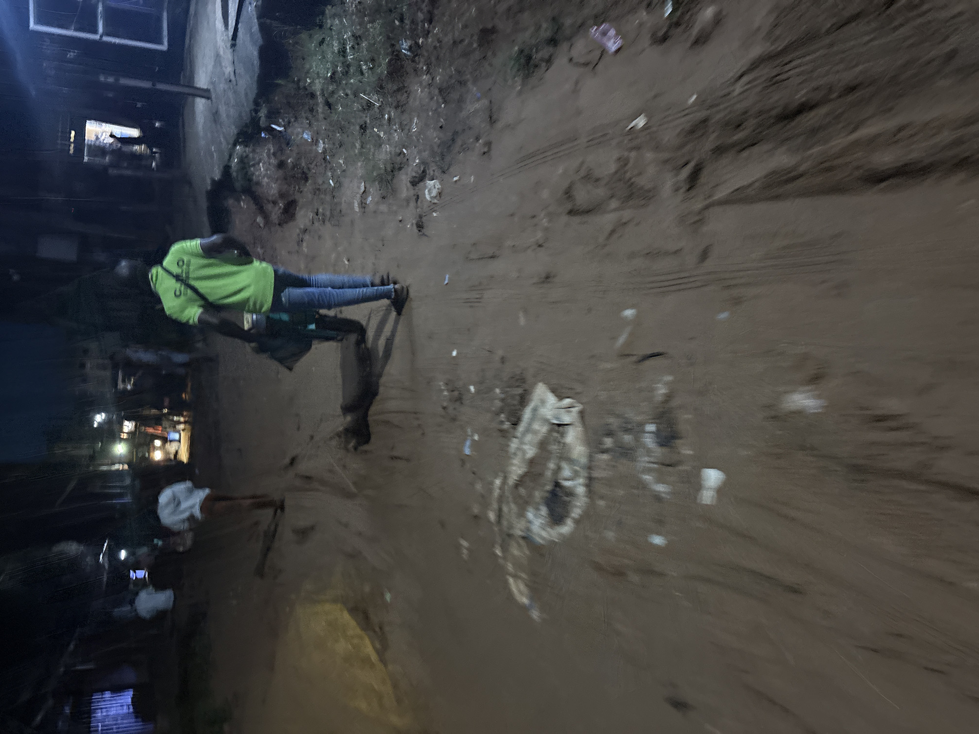 A night scene in Onitsha, Nigeria, shows a person in a bright green t-shirt and jeans pushing a cart on a muddy, unpaved road. The road is littered with debris. In the background, several other figures and structures are dimly visible, including what appears to be a vehicle and buildings. The overall lighting suggests it is nighttime or very early morning.