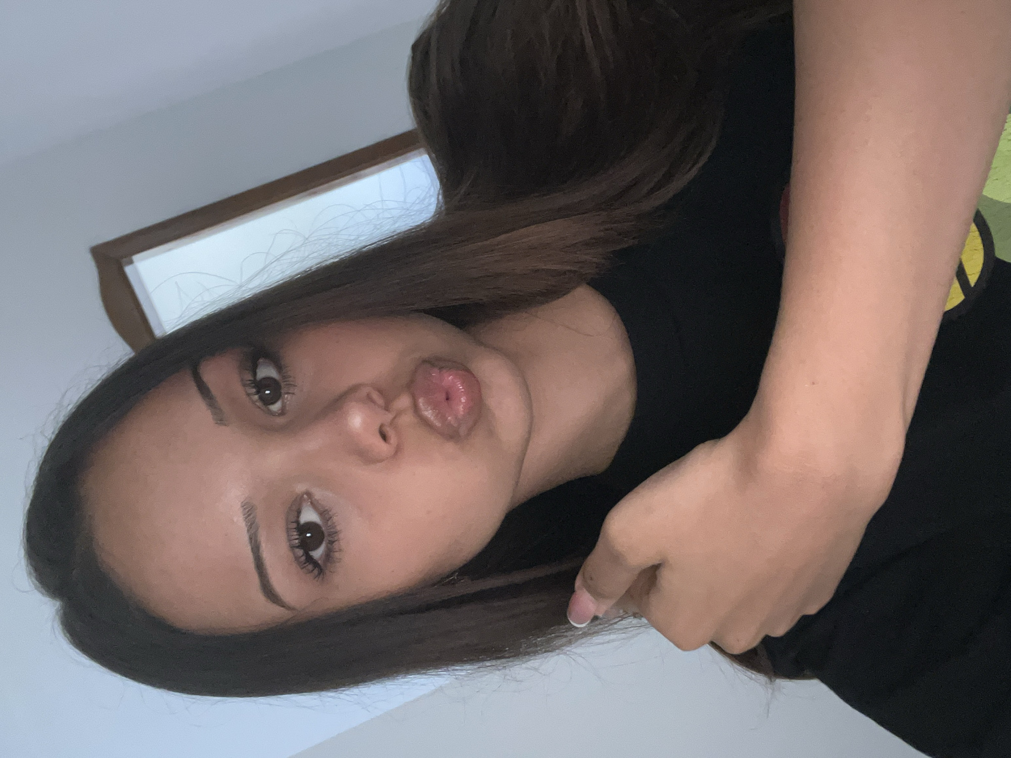 A woman with brown hair and light skin is seen in a close-up shot. Her lips are puckered. She is wearing a black top. The background includes a white wall and part of a window with a wooden frame.
Location: Unknown, Unknown.