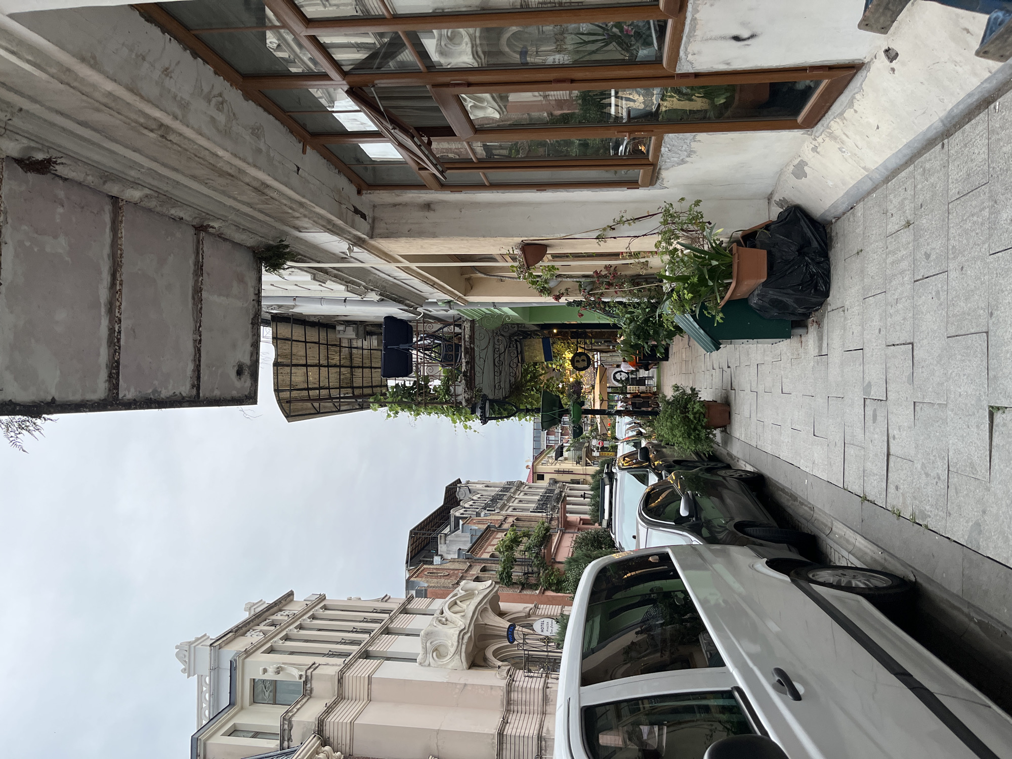 An overcast day in Batumi, Georgia, captures a street scene with vintage architecture and parked cars. The foreground features a white van and other vehicles parked along a stone-tiled sidewalk. Buildings lining the street showcase ornate balconies and windows, with some facades exhibiting signs of age. Lush greenery spills from planters and hangs from balconies, adding a touch of nature to the urban landscape. In the distance, a glimpse of the cityscape reveals more buildings and a few figures walking on the sidewalk. The overall impression is one of a charming, old-world European street.