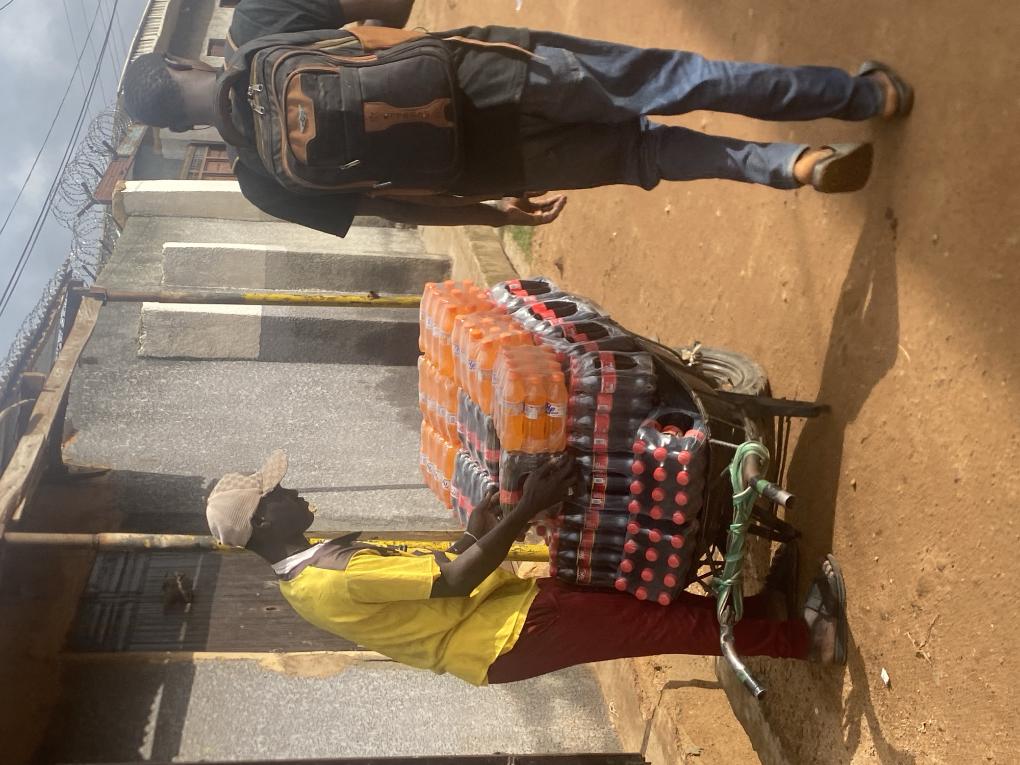 In Bukuru, Nigeria, a man wearing a tan baseball cap and a yellow t-shirt with dark accents is standing next to a wheelbarrow loaded with cases of soft drinks. He is dressed in dark red trousers and appears to be arranging the bottles. The wheelbarrow contains stacks of dark-colored soda bottles, identified by their red caps and branding, and on top, several cases of orange-colored soda bottles.
Another individual, viewed from behind, is walking away from the scene. This person is wearing a black t-shirt, blue jeans, and a backpack with brown accents. They are also wearing dark sandals.
The environment suggests an outdoor urban setting. Behind the man with the wheelbarrow is a building with a textured, light-colored wall and a wooden door. Further in the background, a fence with barbed wire is visible, along with another building. The ground is covered in reddish-brown dirt, indicative of a dusty or unpaved area. The lighting suggests it is daytime, with shadows cast on the ground.
