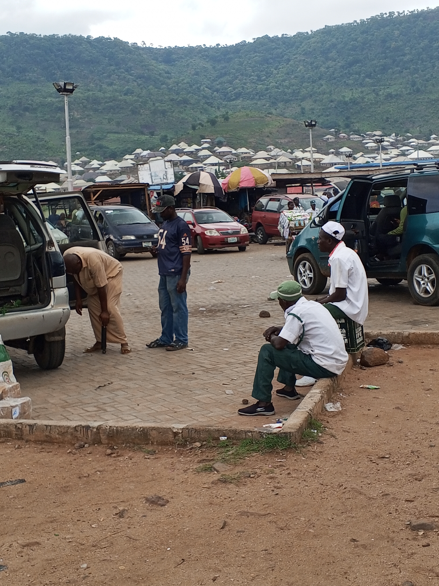 The image depicts a scene in Lokoja, Nigeria. In the foreground, a man dressed in tan clothing is working on a car, while another man in a sports jersey stands nearby. Two men wearing white shirts and hats are sitting on a curb. Several cars and people are in the background. In the distance, a hillside is visible, with houses scattered along its base. The ground is a mix of paved areas and dirt.