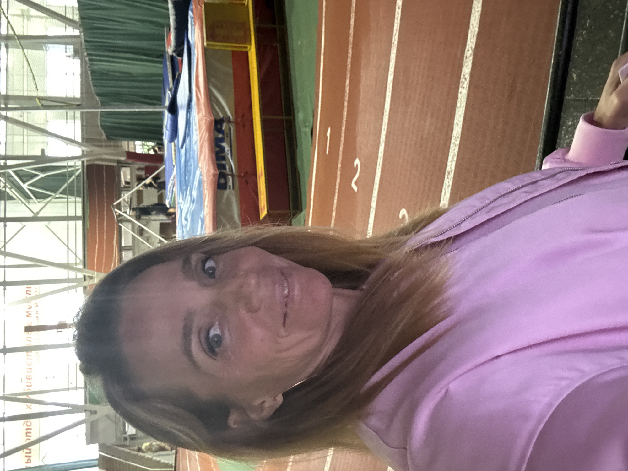 A woman wearing a pink jacket is at an indoor track and field facility. Numbers painted on the track lanes are visible. The location is likely in Saint Petersburg, Russia, given the Cyrillic script visible in the background.