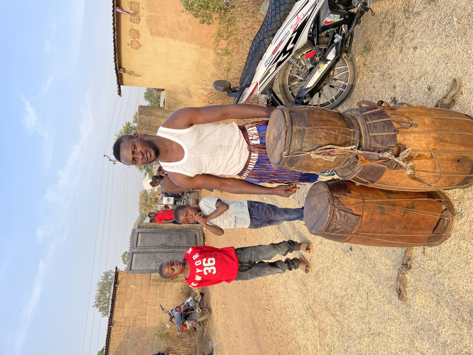 The image captures an outdoor scene, likely in a rural or semi-urban setting, under a clear and bright daytime sky. The ground is dry and dusty.
In the foreground, a group of three individuals is prominently featured, standing near several large, traditional drums. A dark-skinned adult man stands centrally, wearing a white sleeveless tank top. Around his waist, he has a colorful, horizontally striped fabric garment in shades of blue, white, and brown. He looks directly at the camera with a serious or stoic expression.
To his right (viewer's left), a young boy stands next to him, wearing a light blue or grey short-sleeved shirt and dark trousers. His hands are raised near his face or mouth, and he looks towards the man or slightly past him. Further to the left and slightly behind this first boy, another young boy is visible, wearing a bright red short-sleeved t-shirt with white text that reads "NEW YOR 36" (likely "NEW YORK 36"). He also wears dark trousers. Both children are wearing open-toed sandals or are barefoot.
Next to the main man and extending into the mid-ground are three large, traditional-looking drums. They are made of dark wood with animal skin heads, secured by intricate lacing or ropes. These drums suggest a musical or cultural context.
In the mid-ground, a white and black motorcycle is parked, partially visible behind the drums and the main man. Text like "NIKE" and "Smile" is discernible on its body, along with a partial "YAMAHA" logo. Further in the background, a small red motorcycle or moped is also parked.
The background features several simple, single-story buildings constructed from what appears to be mud-brick or adobe, indicating a rustic architectural style. A dark metal gate is visible in the mid-ground. Faint power lines or communication wires are seen against the clear sky in the upper background. Several other people, some wearing red, are visible in the blurred distance, suggesting a gathering or a busy communal area. The strong sunlight casts clear shadows, indicating it is late morning or early afternoon.