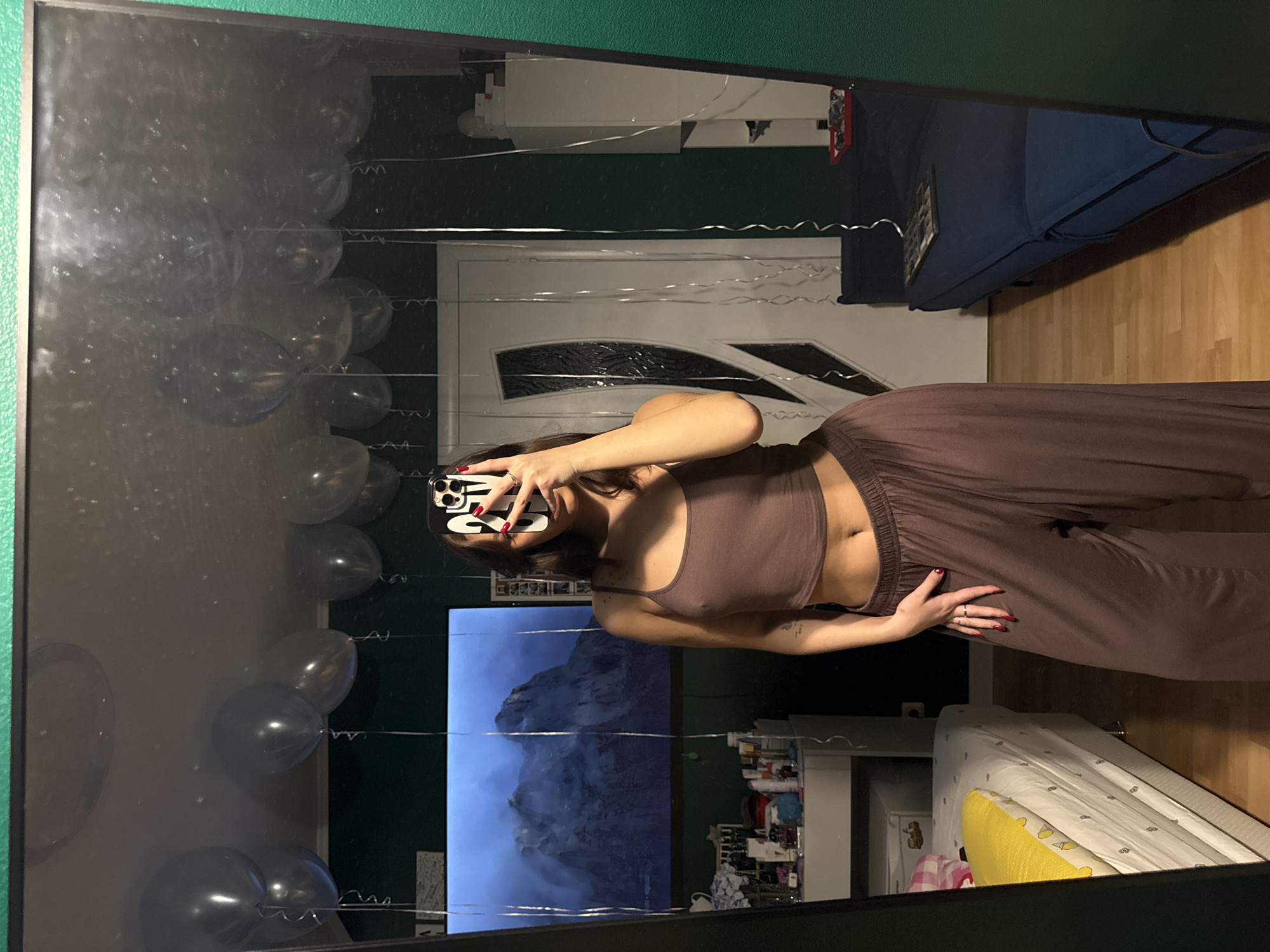 Here is a description of the image:
A woman is taking a selfie in a mirror. She is wearing a brown crop top and matching brown pants. Her phone is covering her face. The background shows a room with decorations, possibly for a party. Visible elements include balloons attached to string, a bed, a white door, and a TV screen displaying a mountain landscape. There is also a glimpse of a shelf filled with various items. The reflections are somewhat distorted due to the curved surface of the mirror.