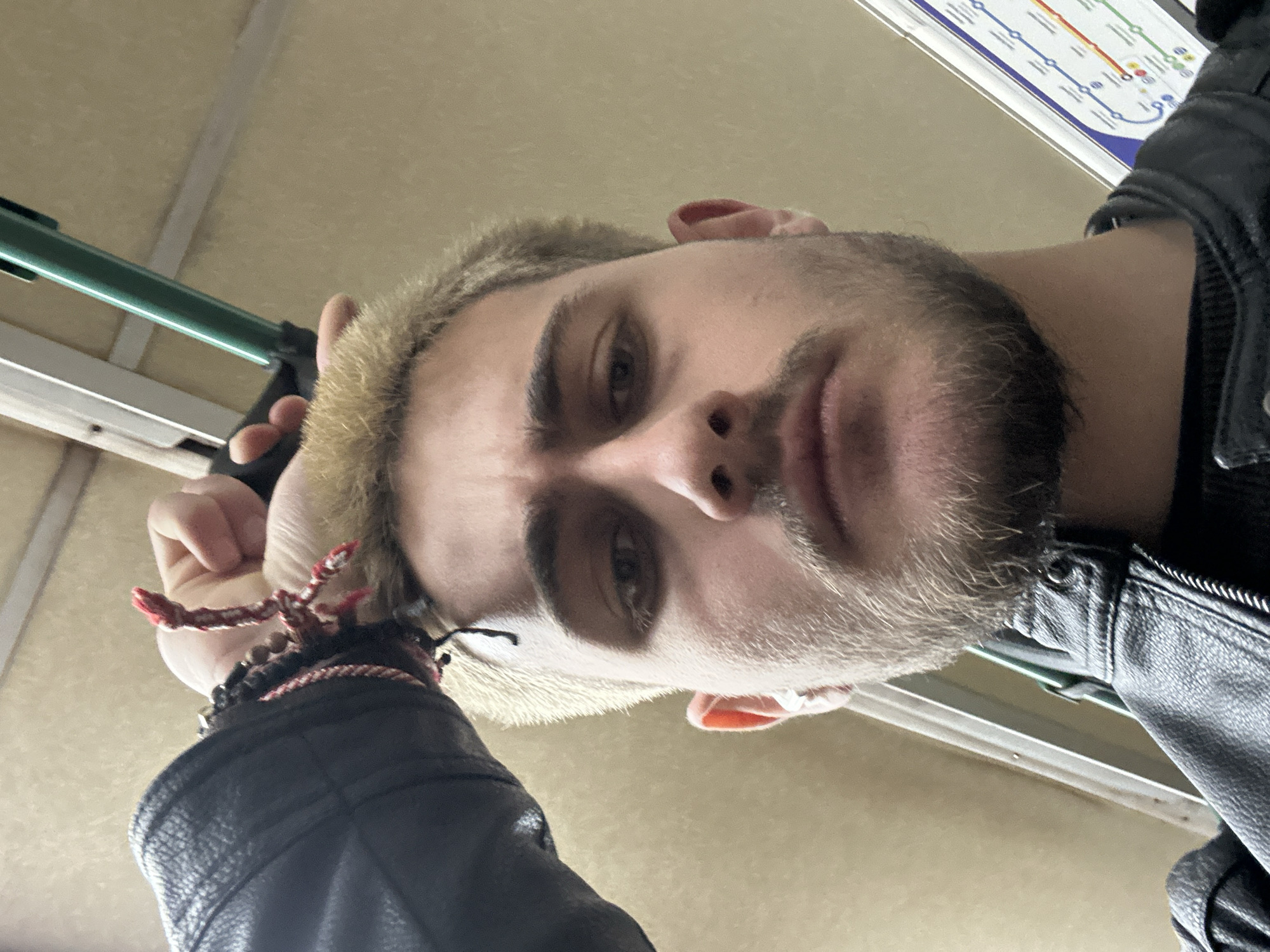 A low-angle selfie of a young man with light brown hair and a beard, wearing a black leather jacket and bracelets, taken on a bus in Sofia, Bulgaria. A map of the Sofia public transport system is visible in the background. The man is holding onto a green handrail.