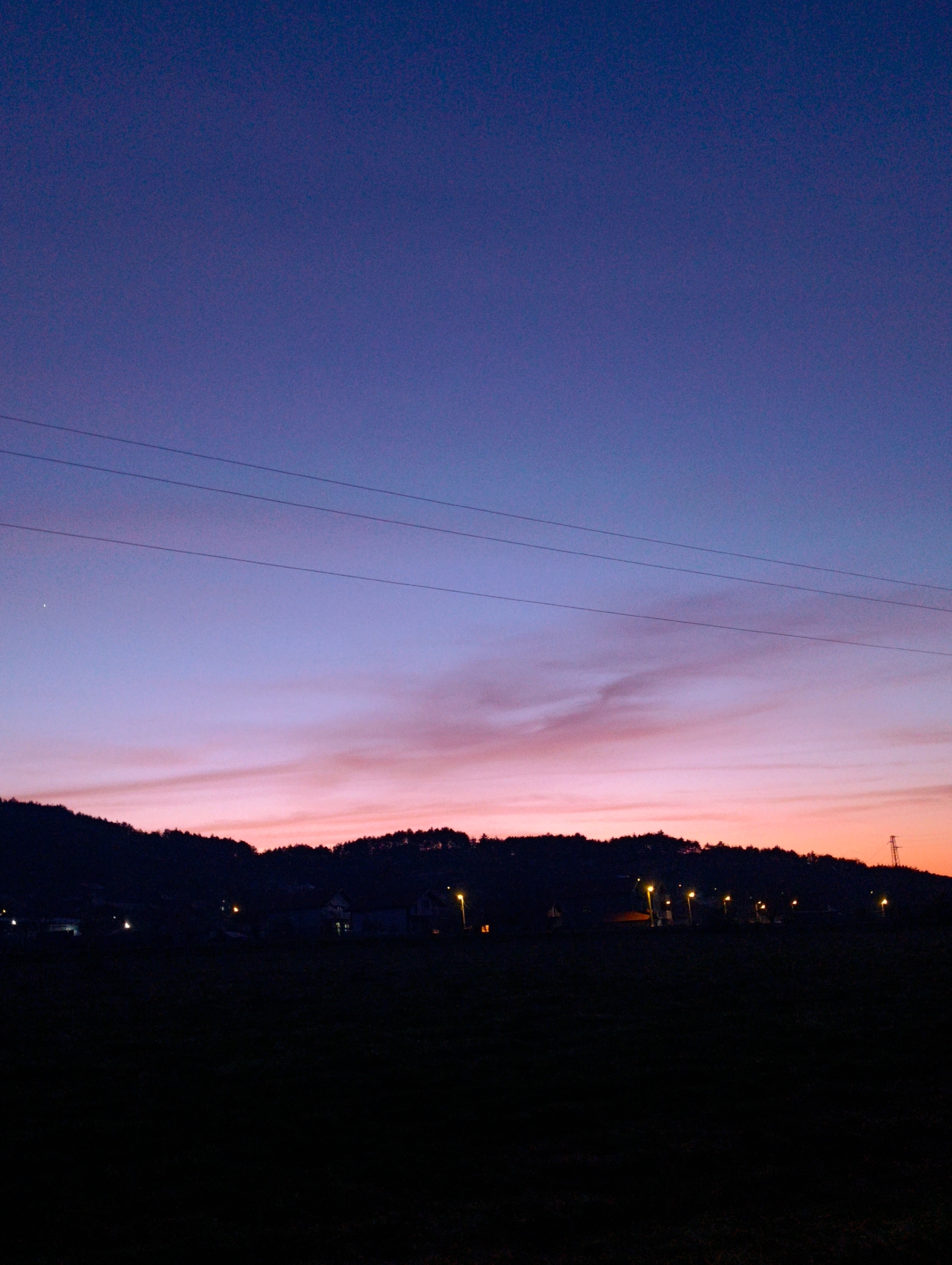 The image depicts a dusk or dawn scene in Shiroko pole, Bulgaria. The sky transitions from a deep blue to a pinkish-orange at the horizon. Three power lines stretch horizontally across the upper portion of the frame. Below the sky, a silhouetted landscape consists of a tree-covered rise, possibly a hill or a small mountain range. Interspersed within this silhouette are points of light, likely from houses and street lamps, indicating a small village or town. A transmission tower stands prominently at the far right of the image. The foreground is completely black, suggesting a field or an unlit area. No people are visible.