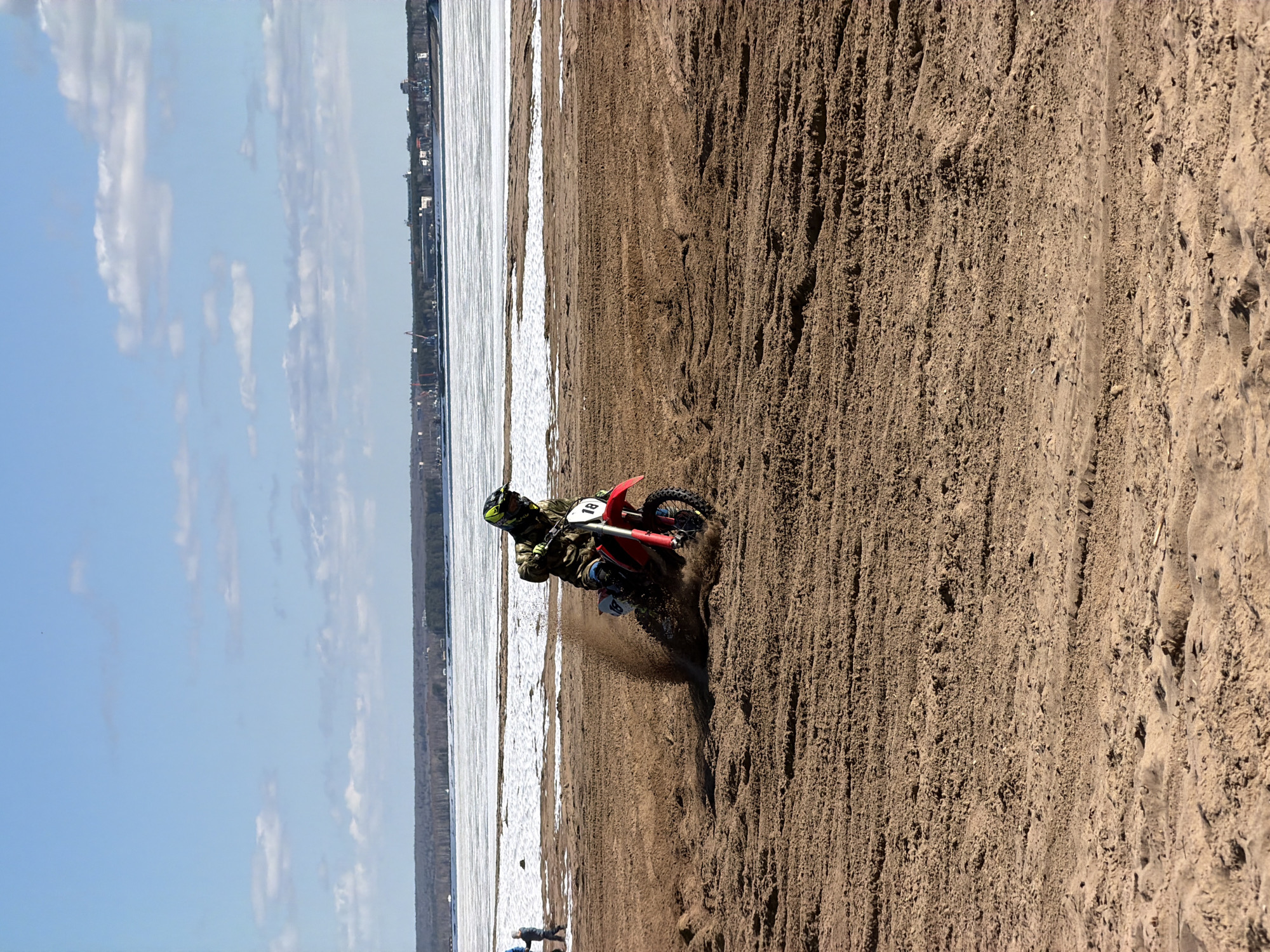 A motorcycle rider is maneuvering a red dirt bike on a sandy beach. The rider is wearing a helmet and protective gear. In the background, a body of water appears to be partially frozen, and the skyline of the city of Chaikovsky, Russia, is visible. The sand appears to be wet and churned by the motorcycle's tires. There are clouds in the sky.