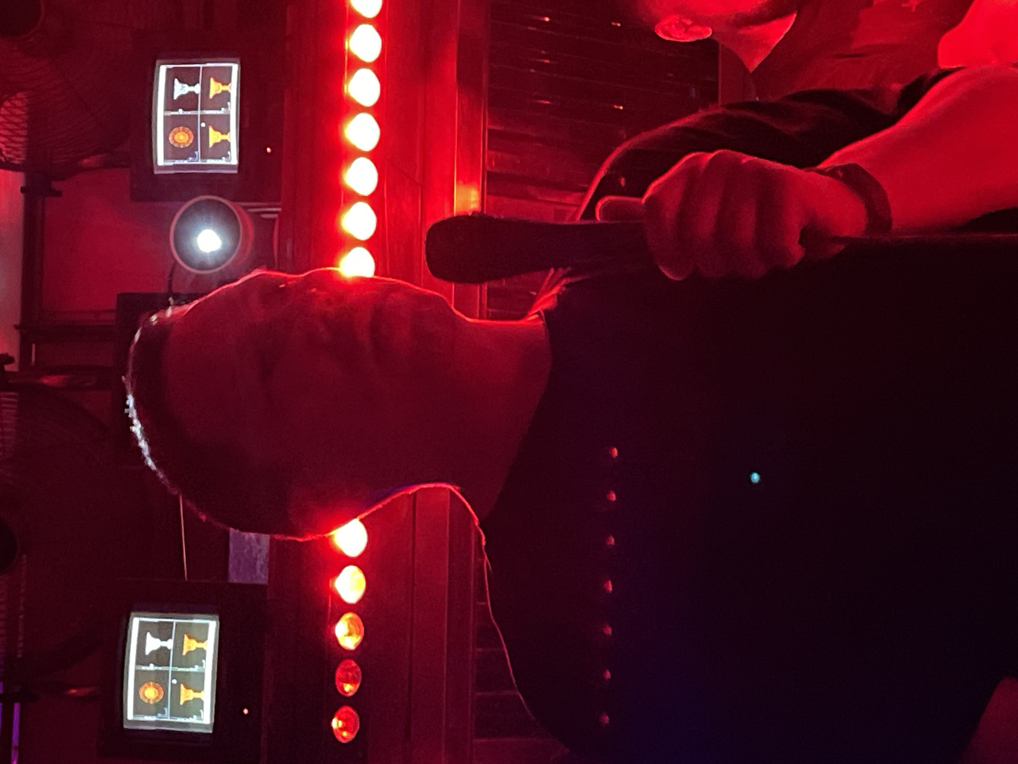 The scene depicts an unidentified indoor entertainment venue, likely a karaoke bar, located within the city of Moscow, Russia. The environment is predominantly illuminated by a strong red light, creating deep shadows.
In the foreground, a male subject is positioned centrally, largely silhouetted against the red lighting. He is facing slightly right, with his head turned towards the right side of the frame. His facial features are obscured. He holds a dark, standard microphone vertically in his right hand, with his thumb gripping it. A dark band is visible on his right wrist. He is wearing a dark, short-sleeved shirt. A partial view of another individual's head and shoulder is visible on the far right edge of the frame, also bathed in red light.
In the background, above a horizontal row of multiple glowing red/orange circular lights, two small, square, retro-style monitors are visible. Each monitor displays a four-quadrant graphic. The common symbols across both screens include a golden medallion/coin in the top-left quadrant, a silver/white trophy in the top-right, and golden trophies in the bottom-left and bottom-right quadrants. Between the two monitors, a bright, circular white light source is emitting illumination. A dark ceiling or upper structure is partially visible, with what appears to be part of a large fan blade in the upper right.