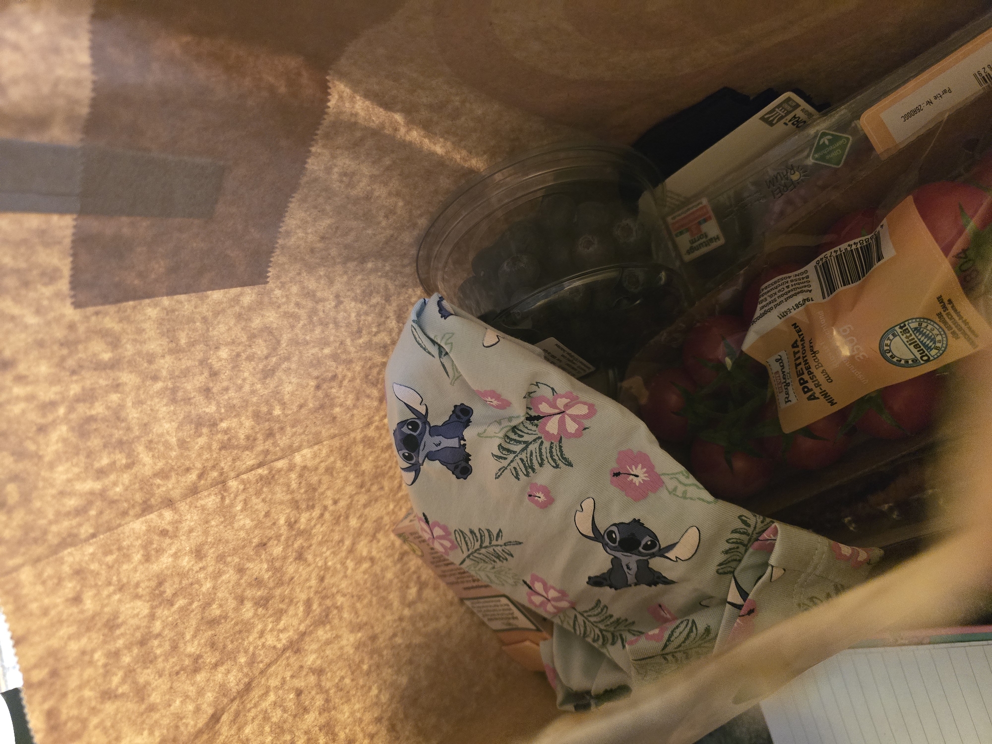 A paper grocery bag contains a carton of blueberries, a package of mini-rispen tomatoes labeled "Appetita" from Gemüsebau Steiner in Kirchweidach, Bavaria, Germany (GGN: 40528528), a container of organic greens (label obscured), and a light blue t-shirt patterned with Stitch characters and hibiscus flowers.
