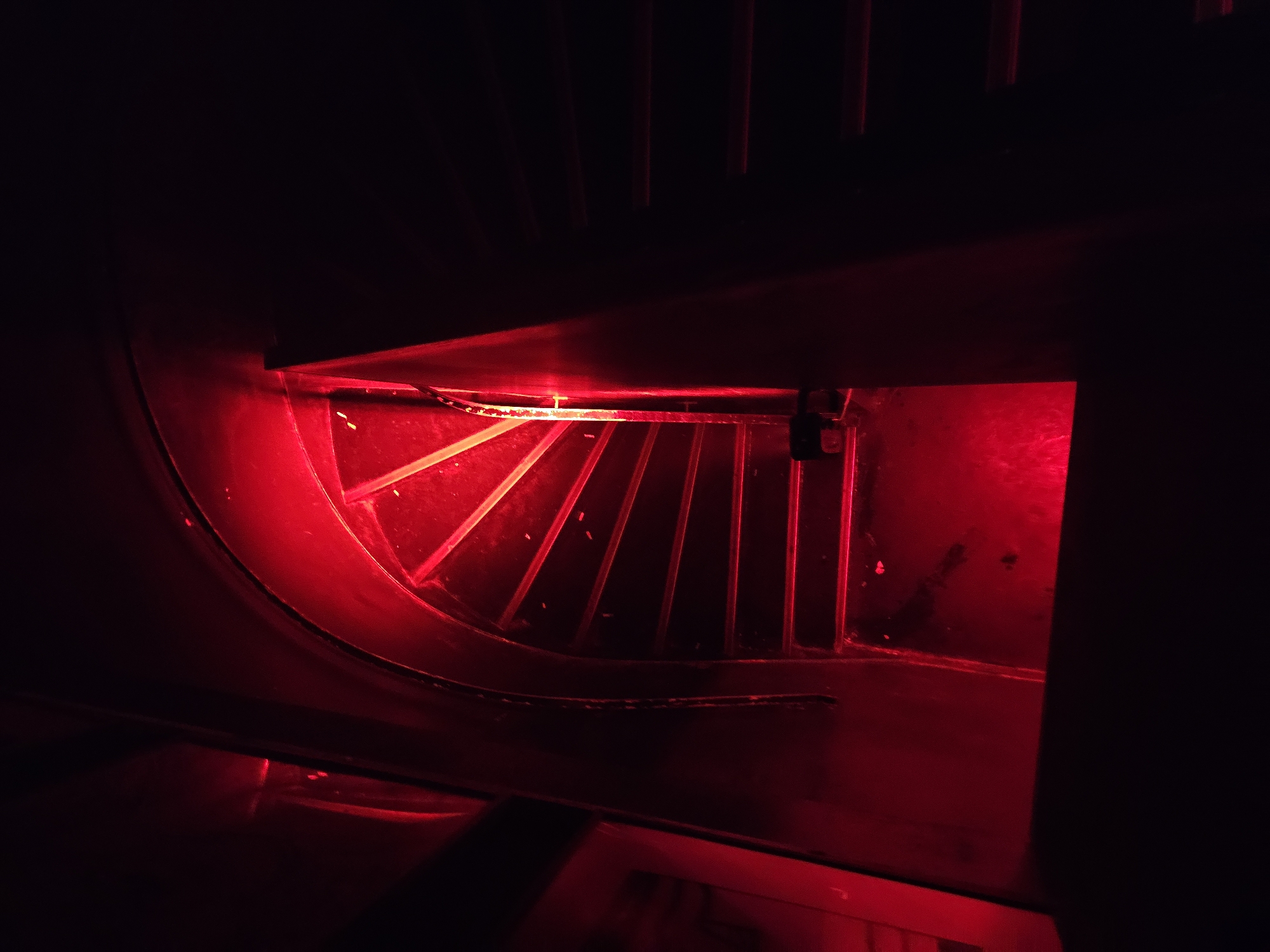 A dimly lit, red-tinted view looking down on a curved grate, potentially inside a piece of machinery or equipment. The location is unknown. The scene is heavily dominated by shadows and the red light source, obscuring details. The grate is composed of parallel bars, and there is a dark rectangular object suspended above it, possibly a sensor or electronic component. The surrounding environment appears to be metallic or industrial. There are no people visible.