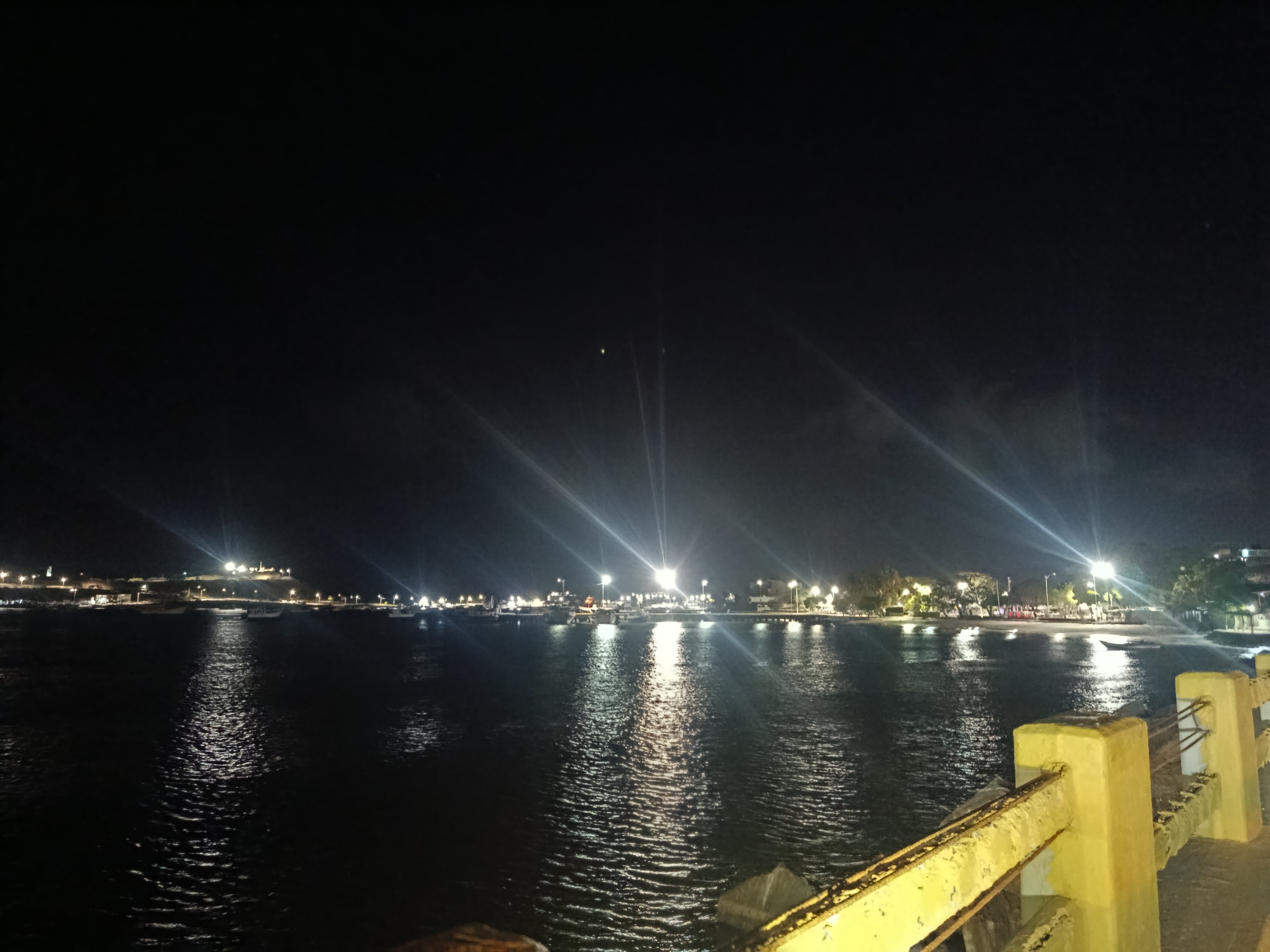 The image captures a night scene in Juan Griego, Venezuela, likely taken from a pier or bridge. The foreground shows a yellow guardrail. Beyond the guardrail is water reflecting the lights of the town in the distance. A dark sky is above the scene with some visible lights from the city. The town's shoreline is illuminated with streetlights and other lights. Several boats are visible on the water. The water's surface is textured with ripples.