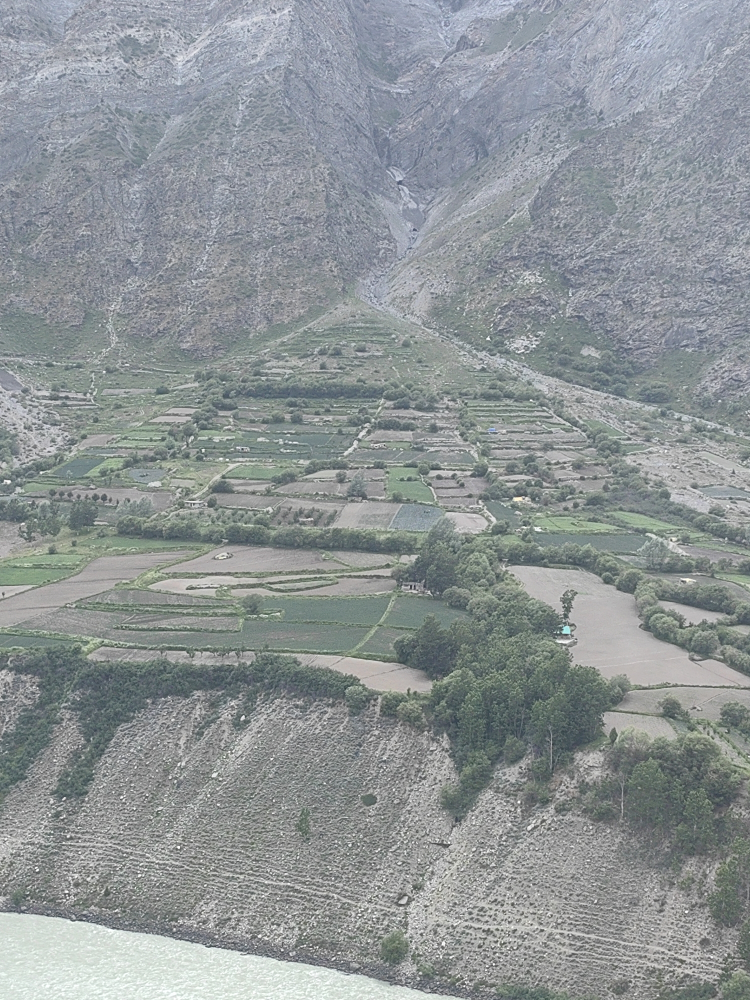 The image depicts a panoramic view of a valley in Tandi, India, characterized by terraced fields and mountainous terrain. The landscape is dominated by rugged, rocky mountains that rise steeply in the background. The slopes of these mountains are partially covered with sparse vegetation and appear to have water erosion channels.
In the middle ground, a series of agricultural terraces are visible, carved into the mountainside. These fields are a mix of cultivated plots with varying shades of green and brown, suggesting different crops or stages of growth. Some fields are irregularly shaped, following the contours of the land, while others are more rectangular. Several small structures, possibly homes or sheds, are scattered throughout the terraced area. A dirt road or path winds through the fields, connecting different sections. A cluster of trees, dense and green, is located on the right side of the terraced area.
The foreground of the image shows a steep, rocky embankment that slopes down to a body of water. The embankment is covered with scattered rocks, dry vegetation, and some patches of green scrub. The water, likely a river or lake, is a muted greenish-blue color and appears to be flowing, with ripples visible on its surface. The overall impression is one of a remote, agricultural settlement nestled within a dramatic, natural environment. There are no people visible in the image.