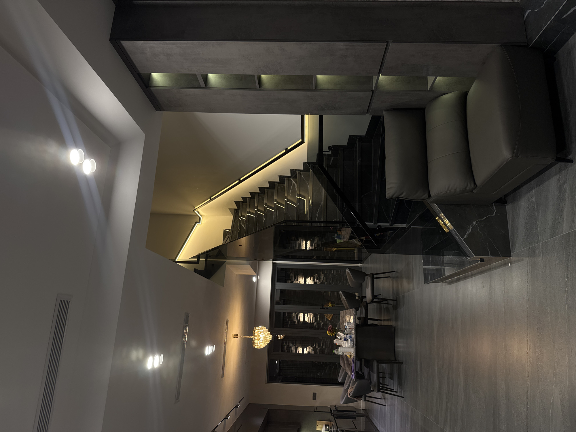 The image depicts the interior of a modern home, seemingly in an urban setting. The home features a prominent staircase with marble steps and glass railings, illuminated by strip lighting along the edge. Adjacent to the staircase, a gray sectional sofa rests on marble-tiled flooring. Above, a dark-colored, modular shelf unit is installed on the wall.
The home's dining area, visible beyond the staircase, includes a long table with chairs arranged around it. A chandelier hangs above the table, providing illumination. The space is characterized by modern decor and muted color palettes, predominantly grays and blacks. The image is taken from a slightly elevated perspective, showcasing the verticality and depth of the interior.