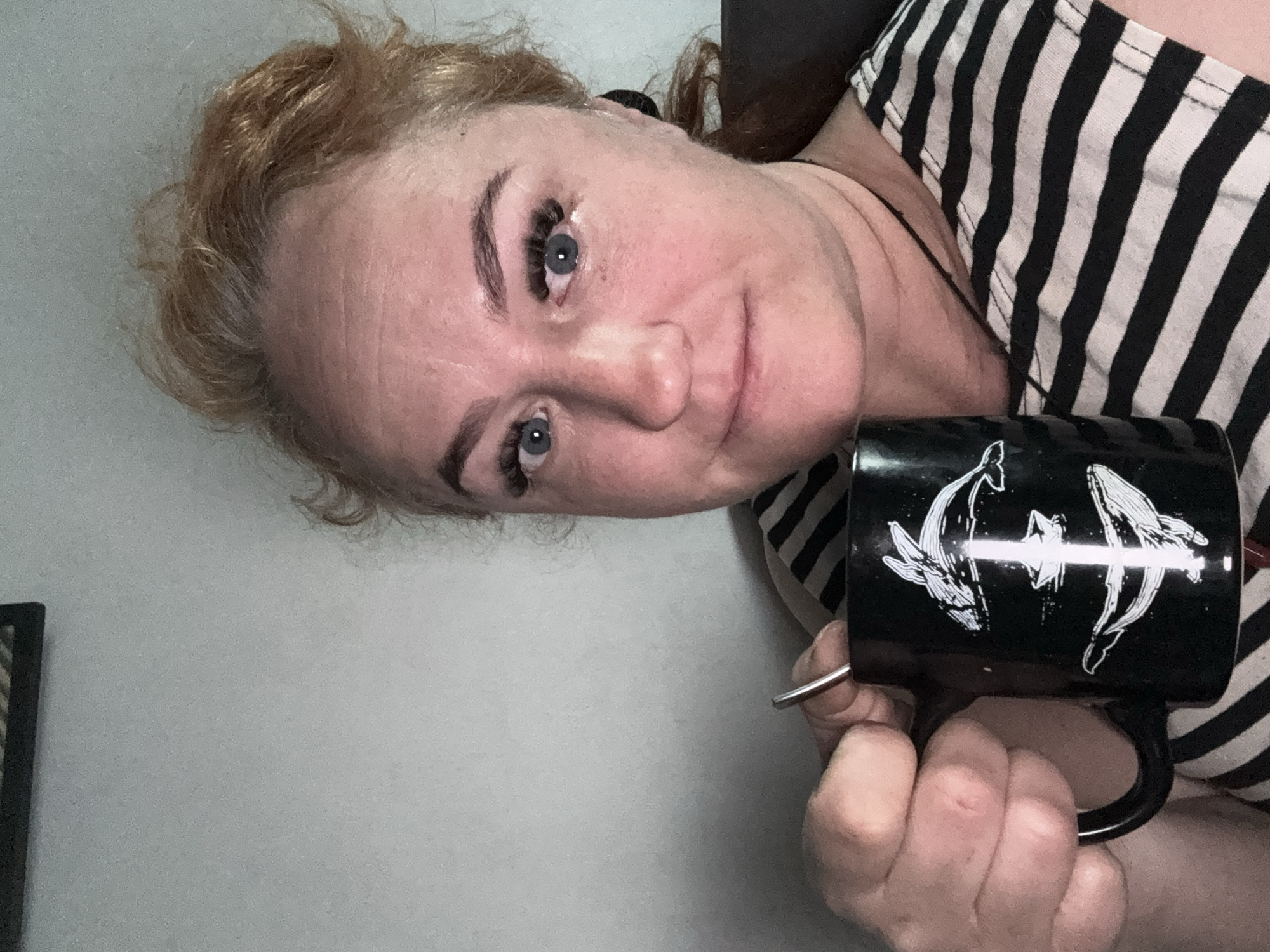 A woman with red hair and blue eyes is holding a black mug with two white whales on it. She is wearing a black and white striped shirt. The background is a light grey wall. The city of Chaikovsky, Russia is not discernible from the image.